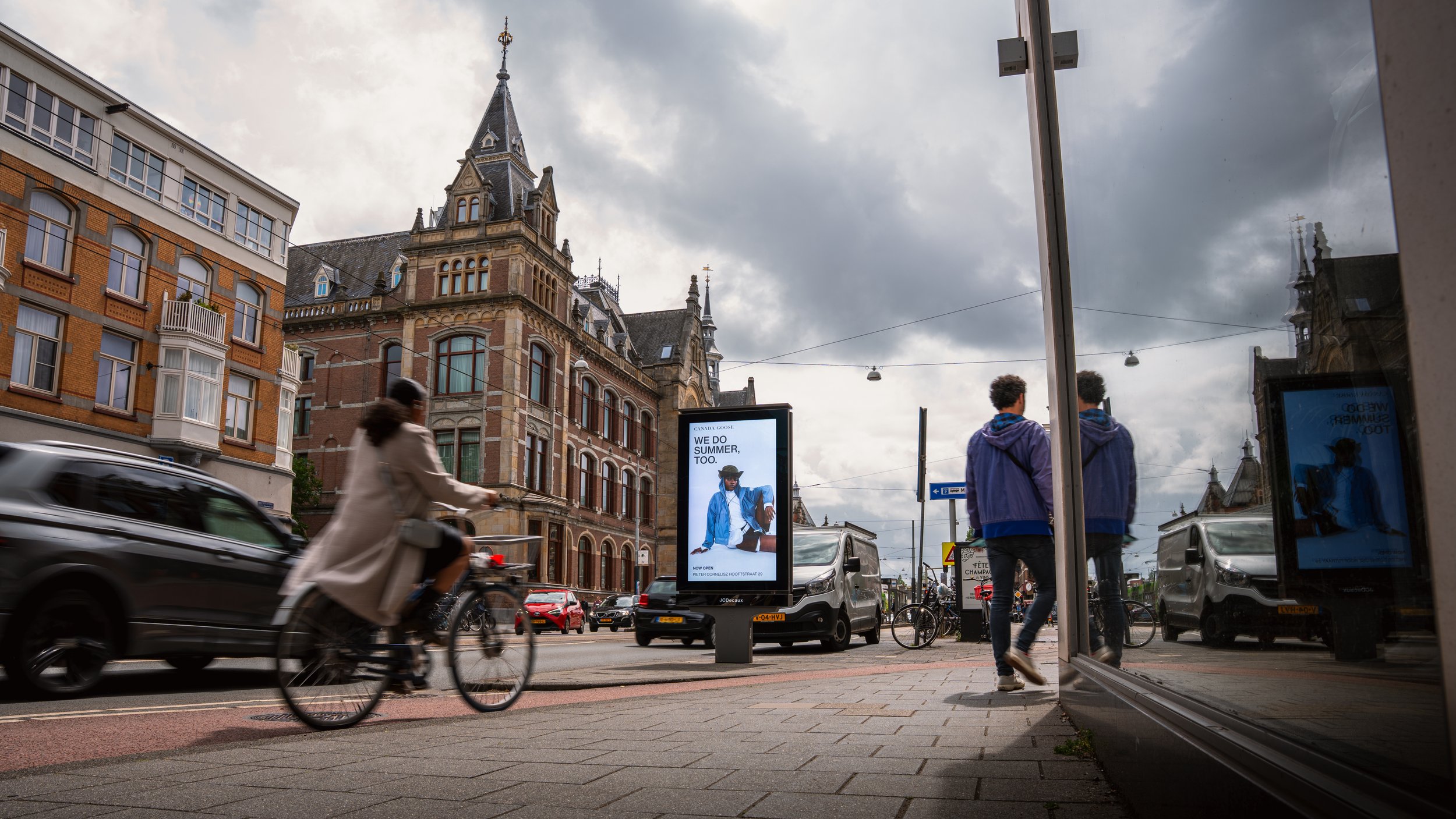Straatbeeld met mensen die lopen en fietsen, auto’s en een digitaal reclamebord met een model en tekst, historische gebouwen, een bewolkte hemel, en een glazen deur die reflecteert op de rechterkant.
