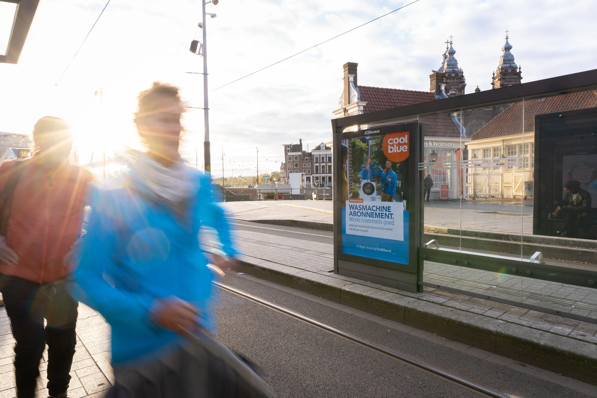 Twee mensen lopen voorbij een bushokje met een poster over wasmachine abonnementen, terwijl de zon ondergaat of opkomt op een stedelijk tram- of treinperron.