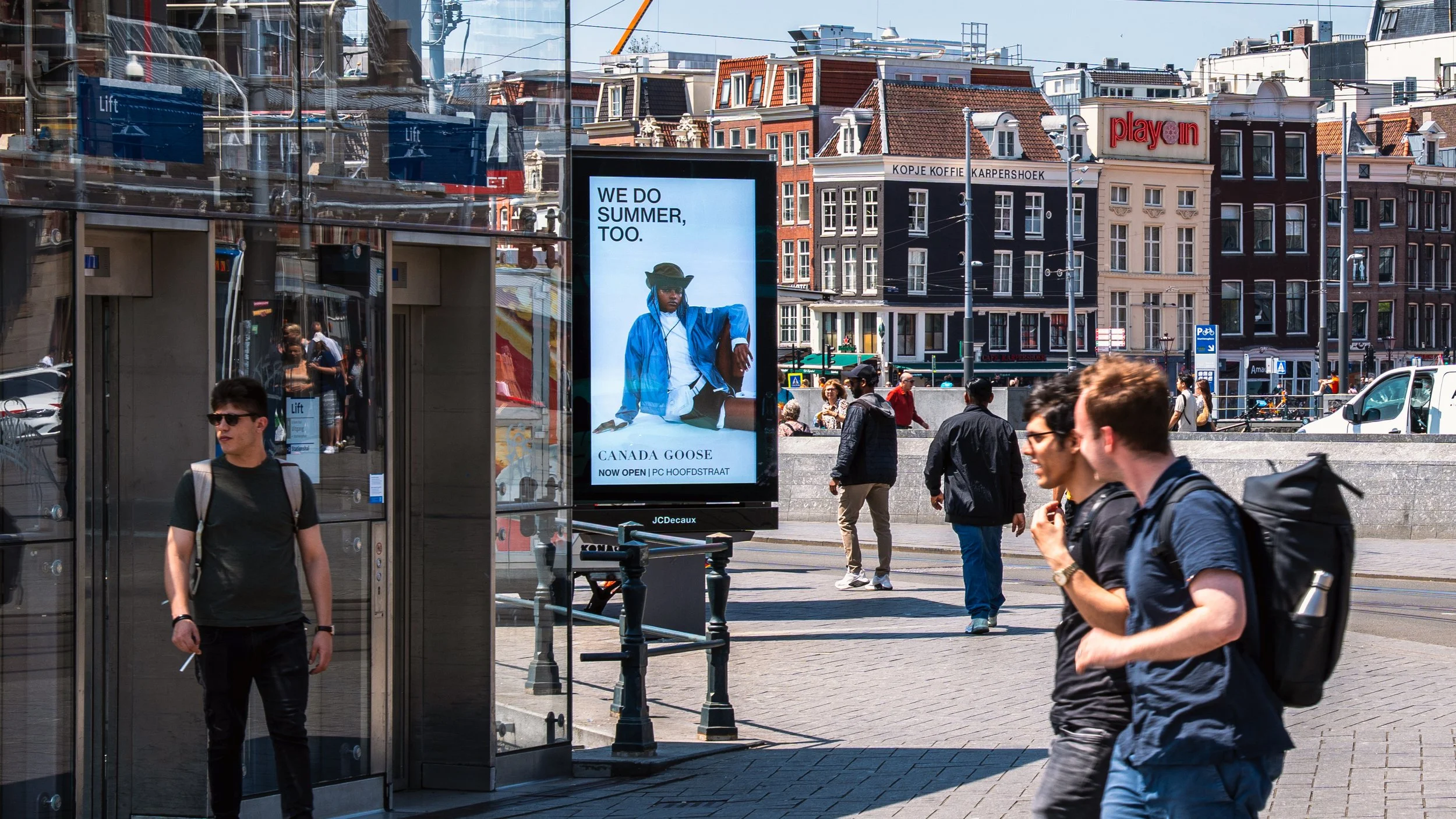 Straatbeeld van Amsterdam met mensen die lopen, een digitale reclamezuil met een advertentie, en rijen kleurrijke oude gebouwen op de achtergrond.