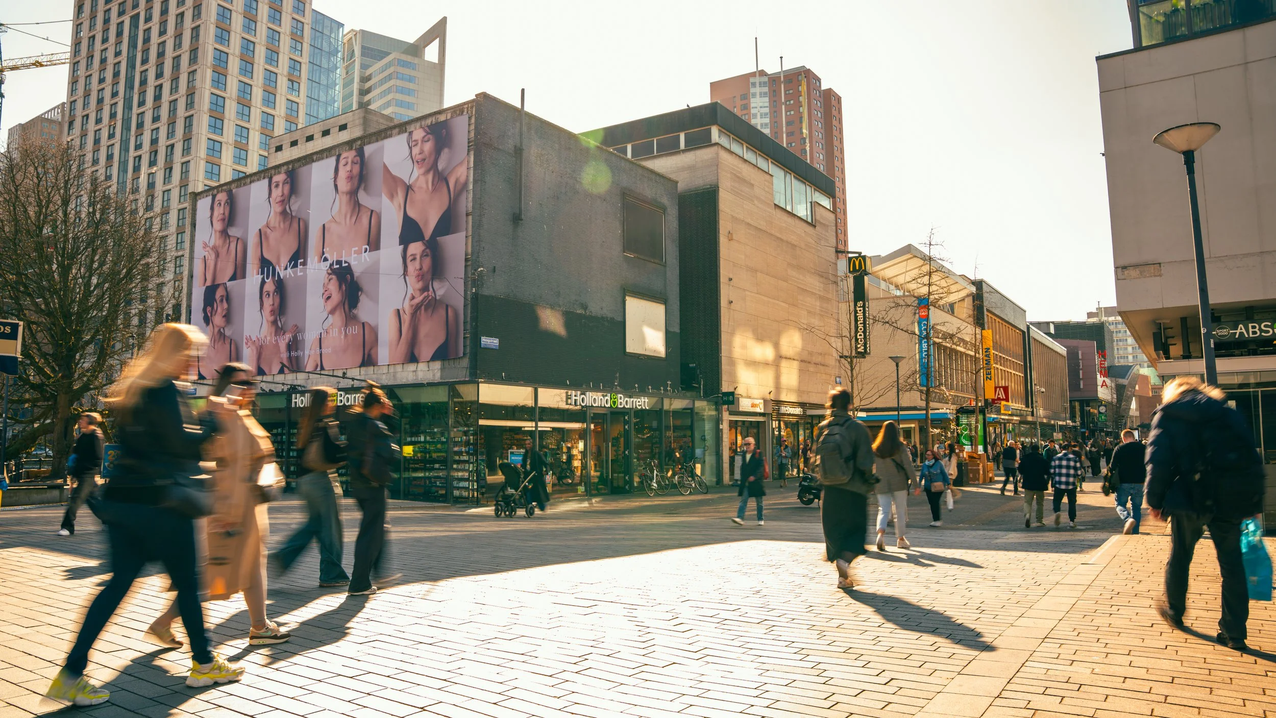 Een druk stadsplein met mensen die lopen, winkels en een groot reclamebord met portretten van een vrouw, en een McDonald's restaurant op de achtergrond.