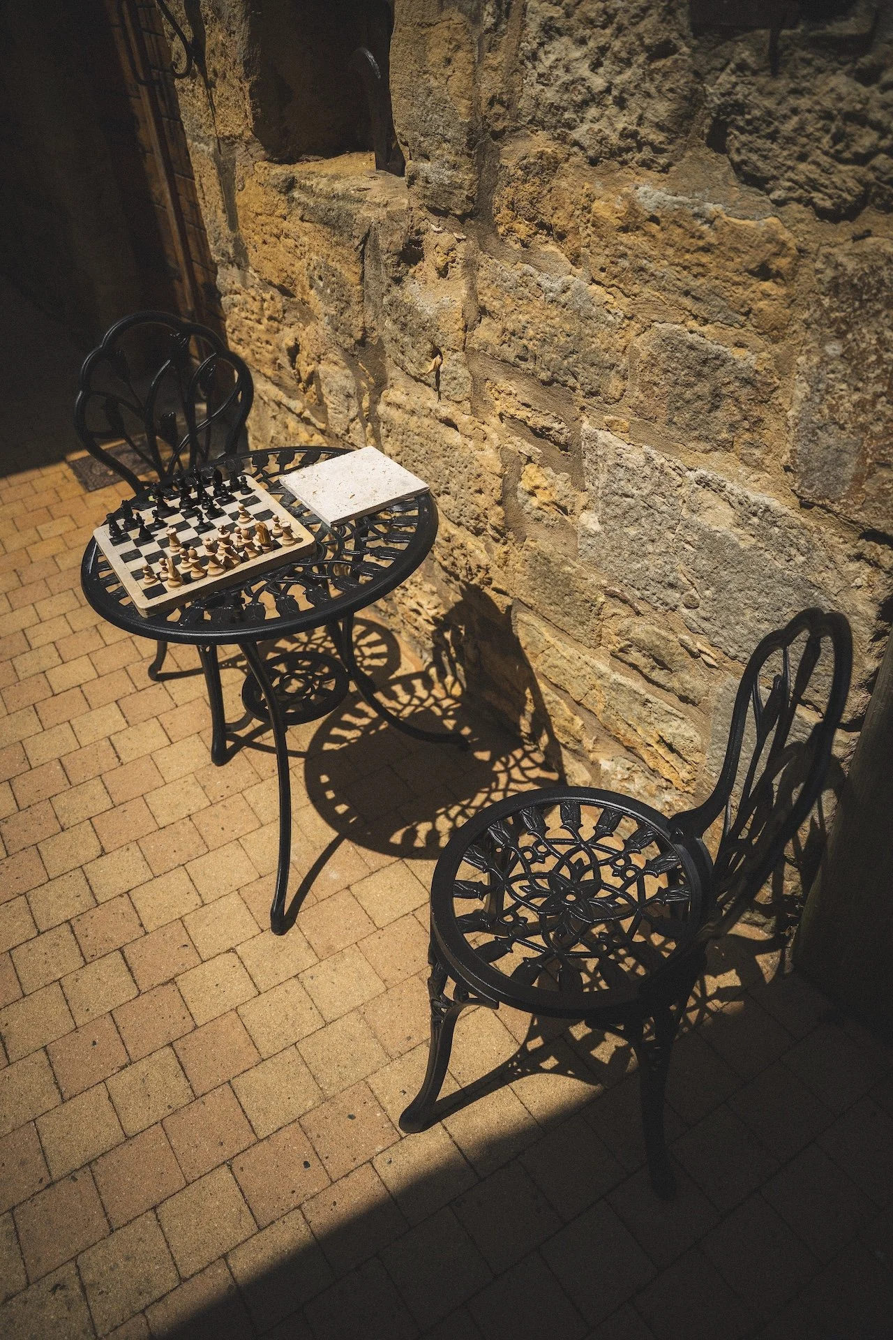 Een zwart ijzeren tafel met een schaakbord en een stuk papier, samen met een zwart ijzeren stoel met een mooi patroon, staan op een stenen vloer naast een bakstenen muur.