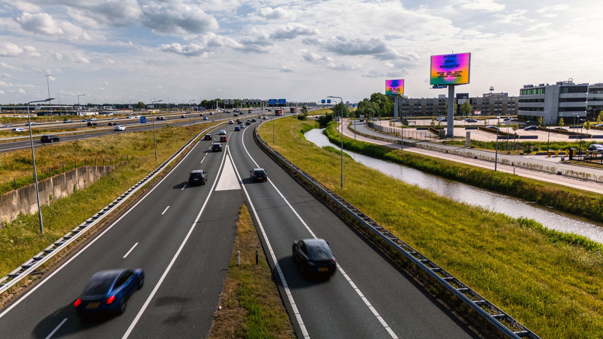 Een snelweg met enkele auto's en een waterpartij langs de weg, enkele digitale reclameborden op de achtergrond en een gebouwencomplex.