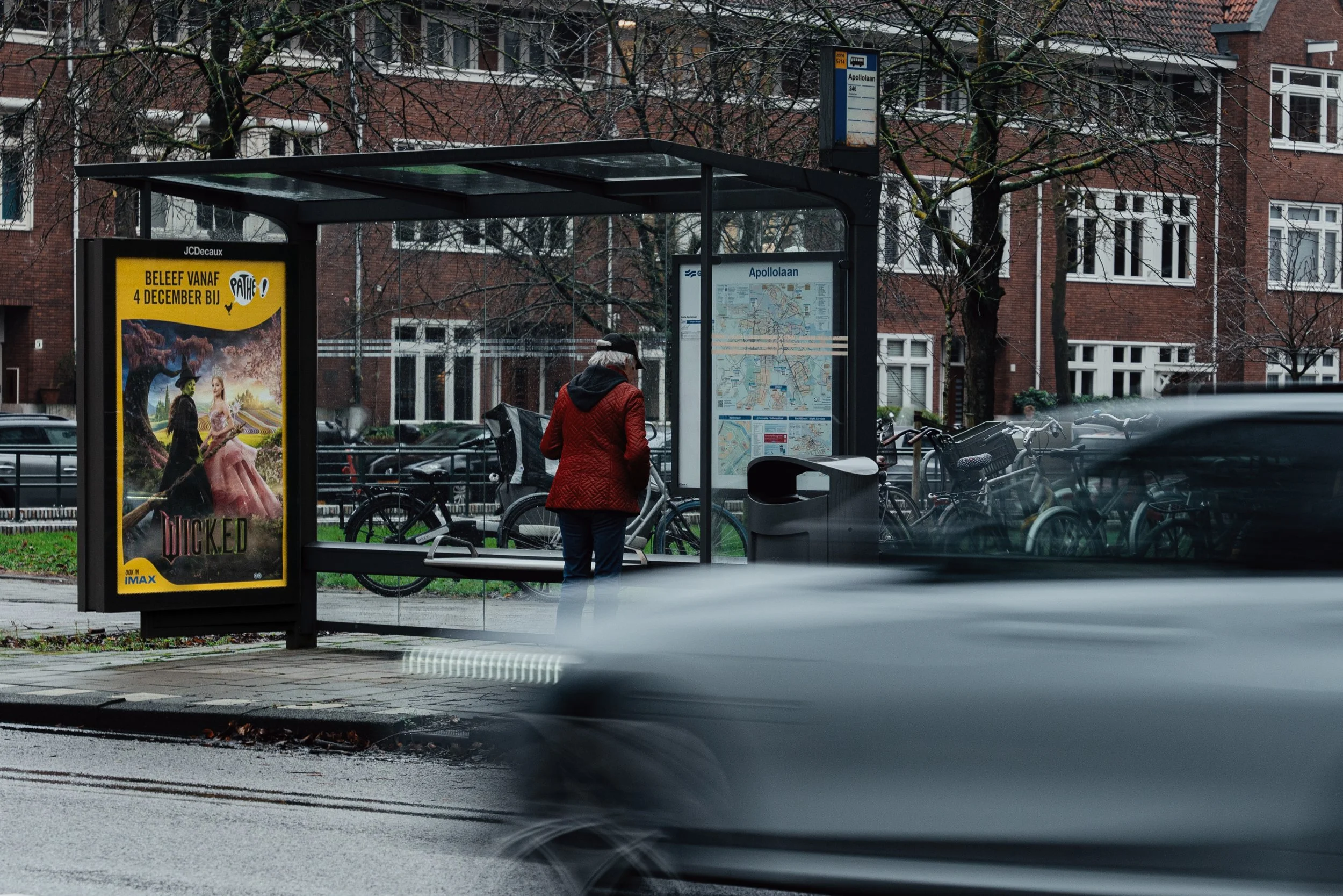Een persoon wacht bij een bushalte met een stadskaart, omgeven door fietsen en een op straat rijden auto, met een advertentie voor een film op de bushalte.