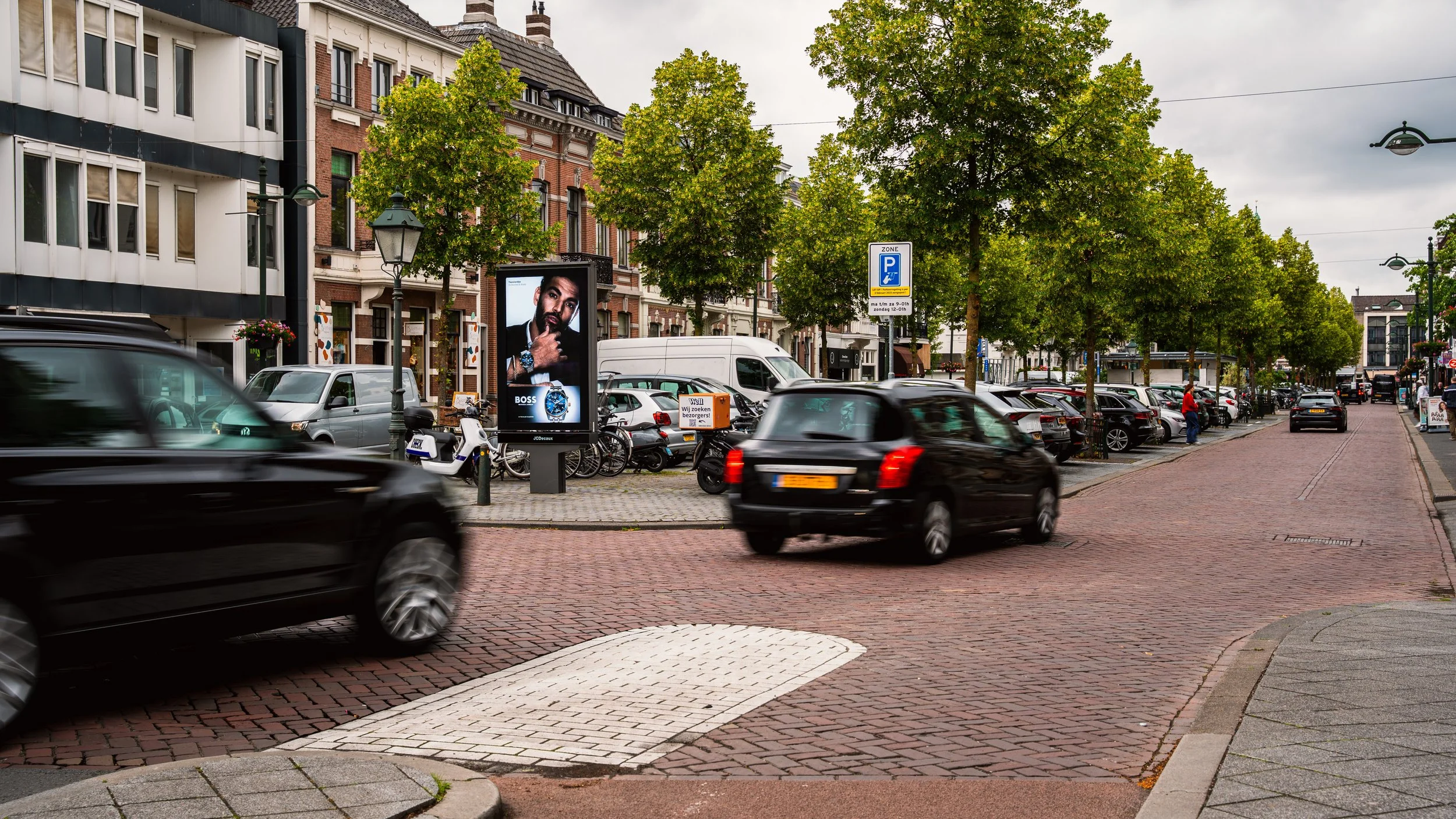 Straat met geparkeerde en rijdende auto's, een digitale billboard met een advertentie, bomen, een ondergrondse parkeergarage en gebouwen op de achtergrond in een stad, bewolkte hemel.