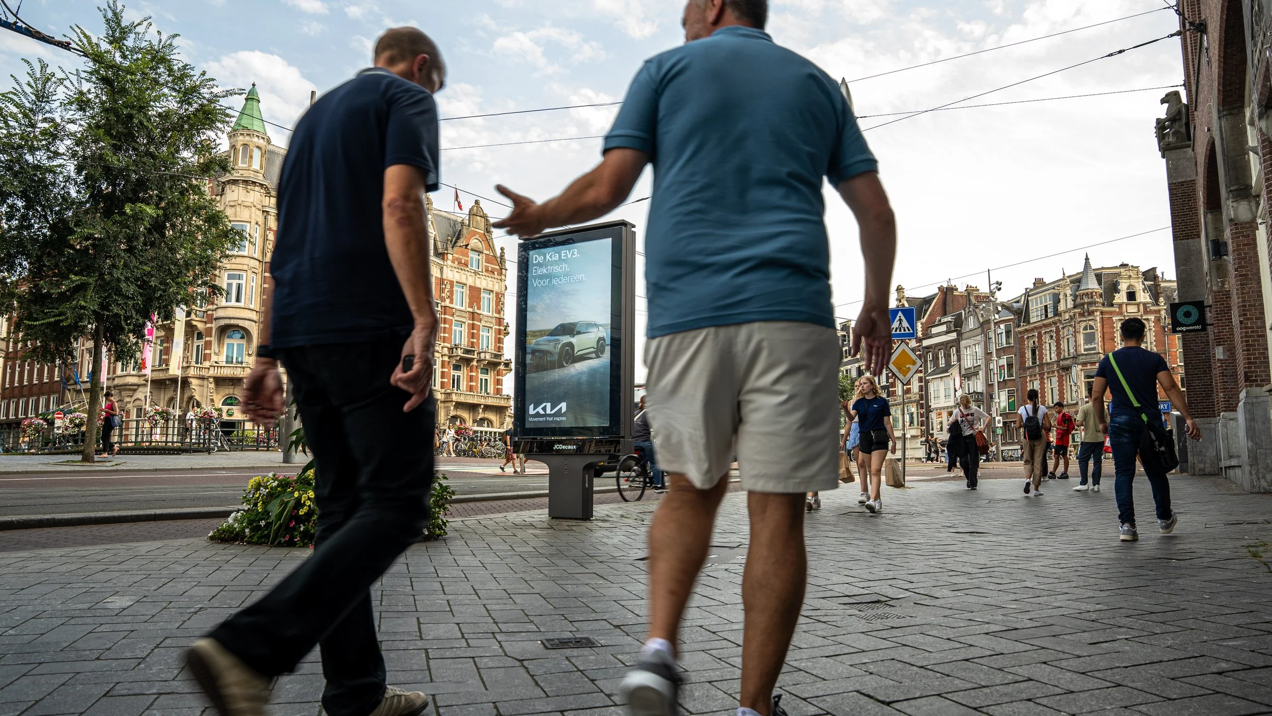 Mensen lopen over een stadsplein met klassieke gebouwen en een digitale advertentiebord.