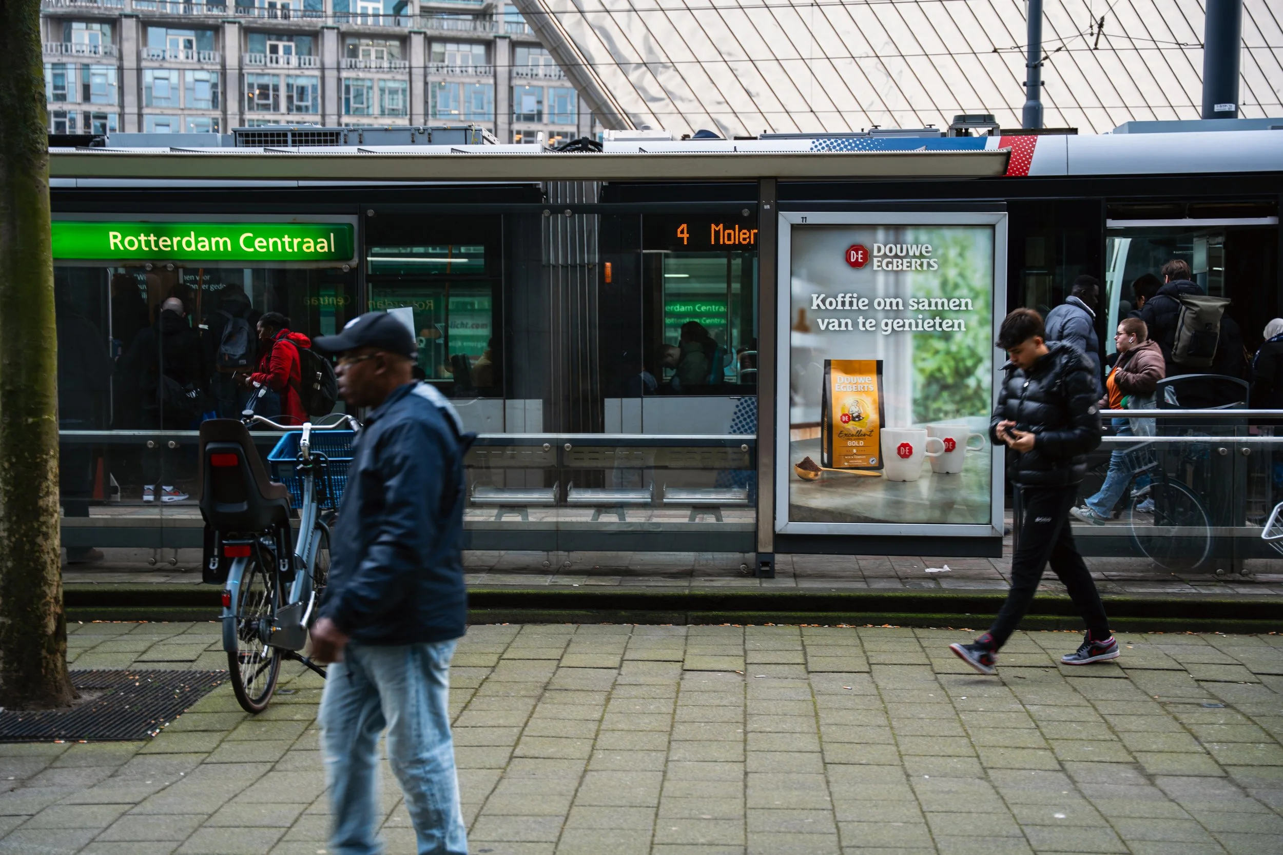 Straatbeeld met een tramhalte in Rotterdam, met mensen die wachten en wandelen, een fietser en een advertentie voor koffie.