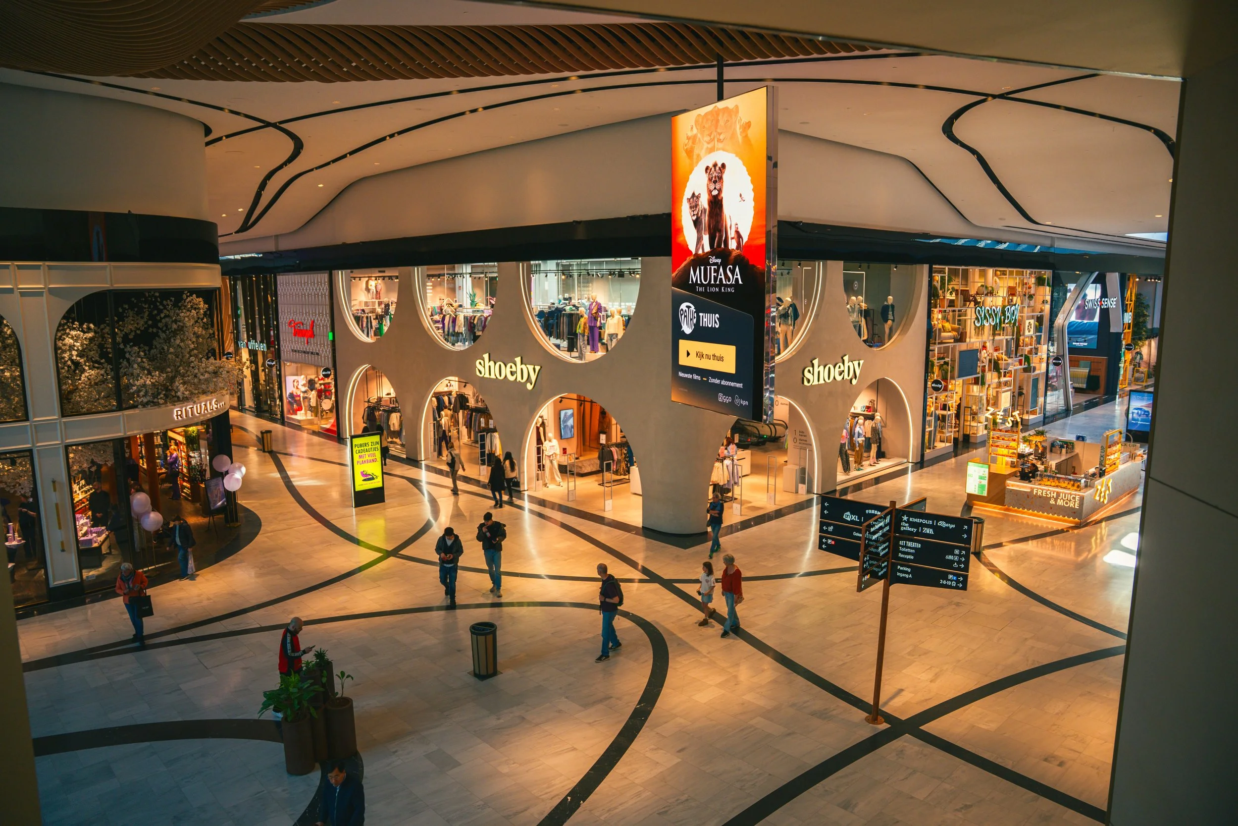 Binnen in een winkelcentrum met winkels als Shoeby en andere winkels op de eerste verdieping, mensen lopen door een grote open ruimte met zwart-witte marmeren vloeren en een digitaal reclamebord.