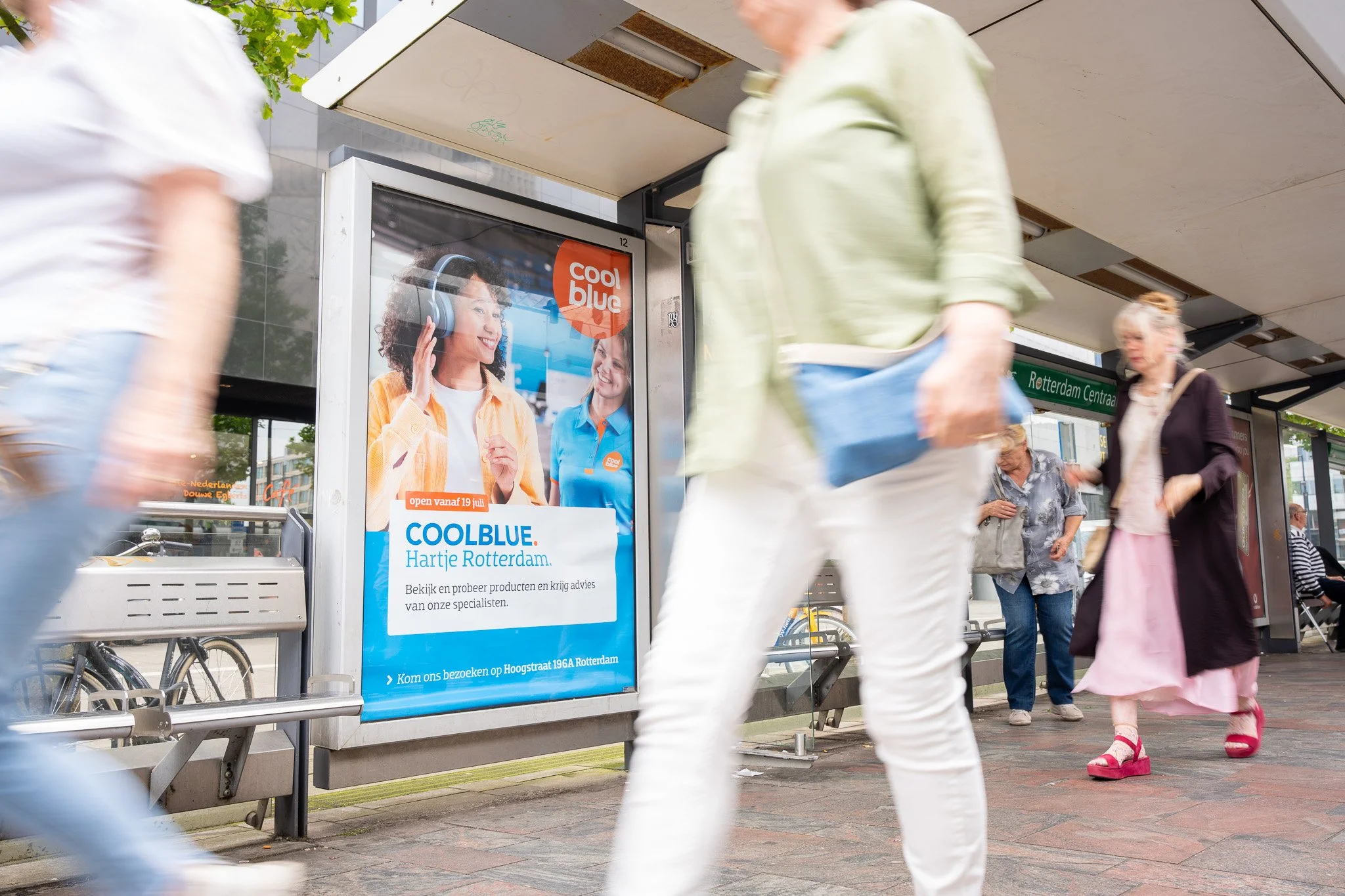 Mensen wachten bij een bushalte in Rotterdam, met een poster op de reclamebuis die reclame maakt voor COOLBLUE.