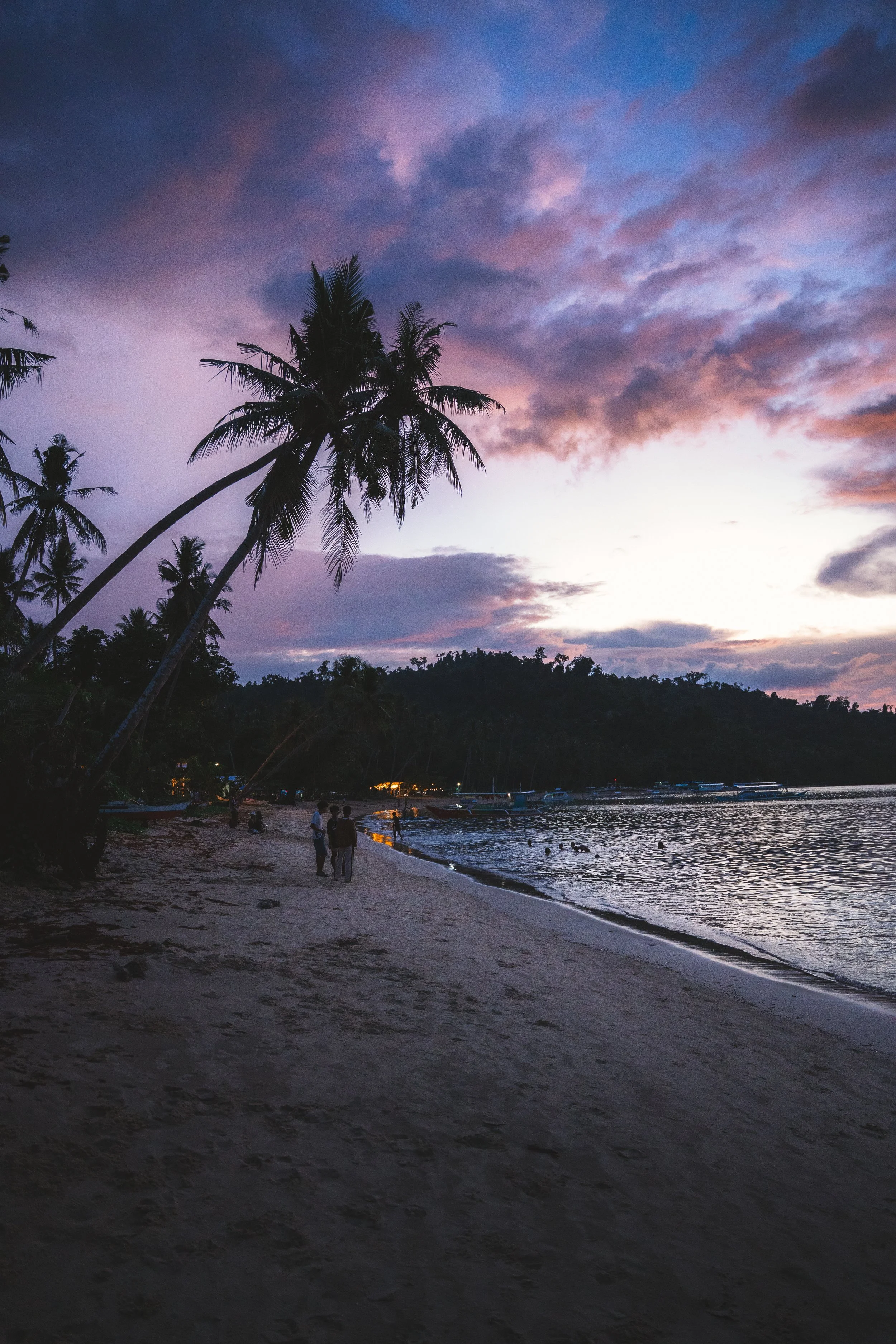 Strand bij zonsondergang met palmbomen, mensen wandelen langs de kust, boten in de zee, bewolkte hemel met roze en blauwe kleuren.