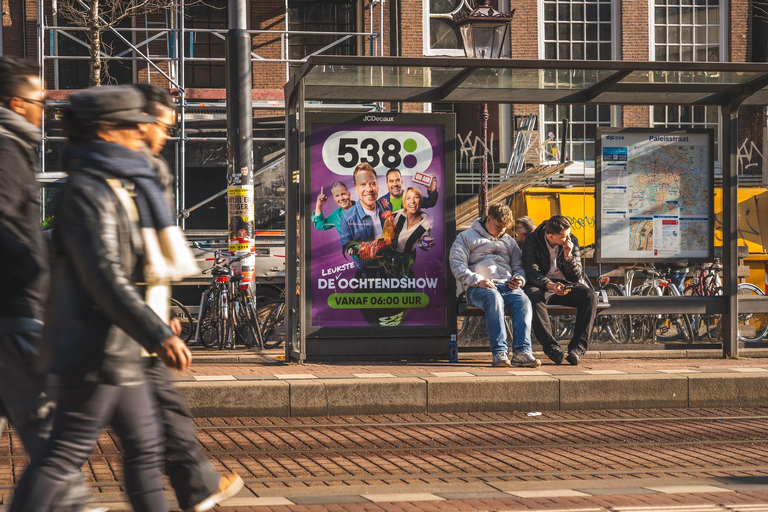 Drie mensen wachten bij een bushokje, twee mensen lopen langs de bushokje. Er staat een advertentie voor '538', met foto's van mensen en de tekst 'De ochtendshow vanaf 06:00 uur'.