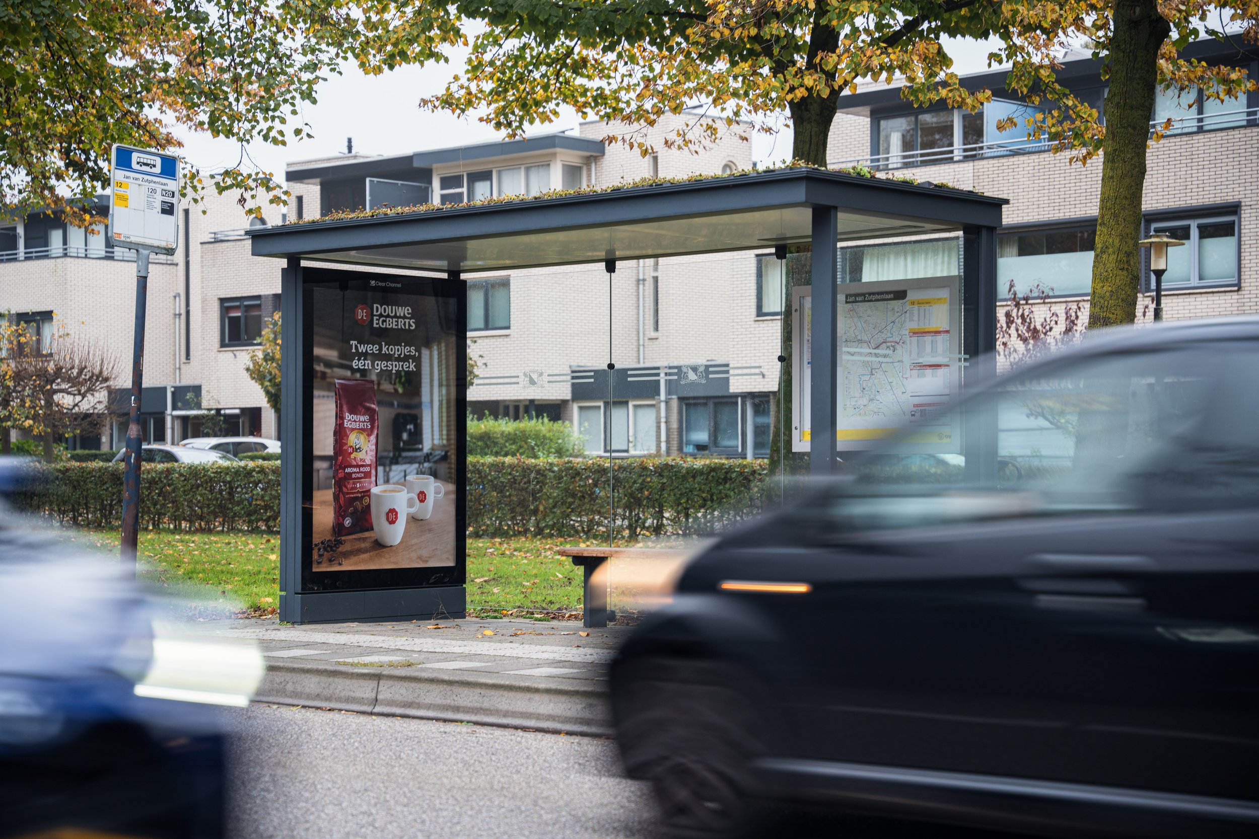 Een bushalte met een advertentie voor Douwe Egberts koffie en een routekaart ernaast, met auto's die voorbijrijden op de voorgrond.