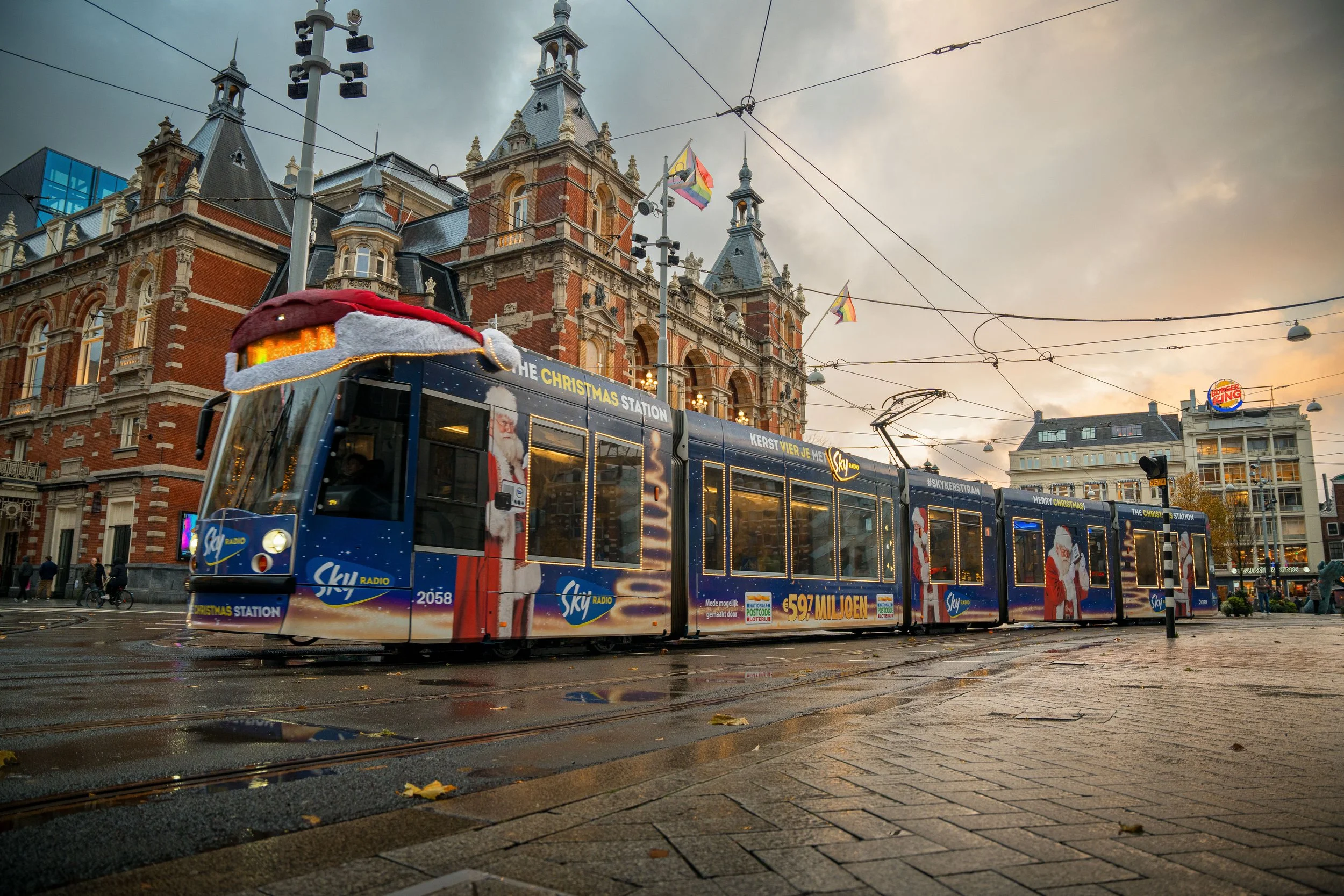 Een stadsbus versierd met een gigantische Santa claus hoed en kerstdecoraties, rijdend op een tramspoor voor een groot historisch gebouw met kerstversiering en vlaggen, tijdens een bewolkte dag.