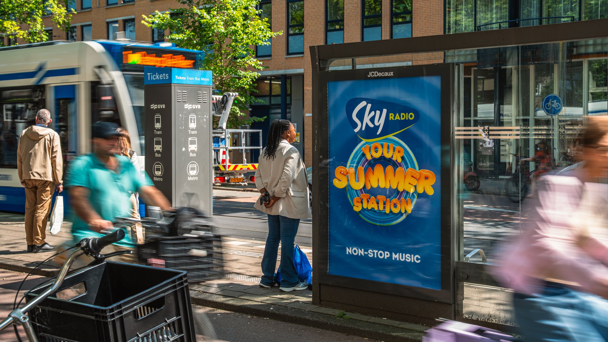 Een bushokje met een Sky Radio campagne voor 'Your Summer Station' met non-stop muziek, nabij een bushalte met mensen, een tram en een fietser in een stedelijk omgeving.