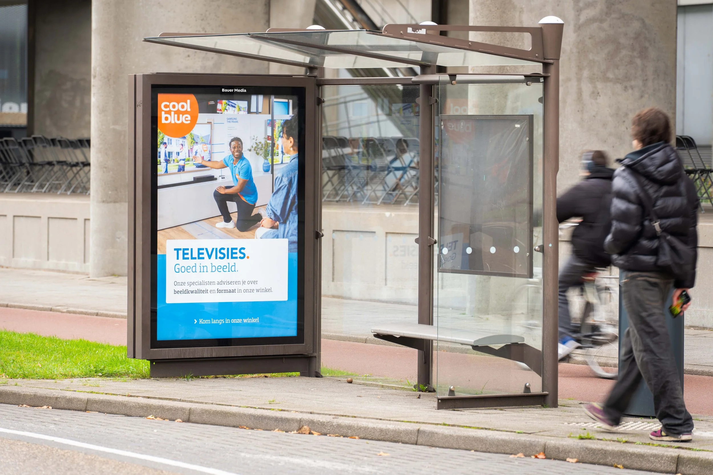 Advertentiebus bij bushokje met digitale display, mensen lopen voorbij, straat, en een groen grasveld.