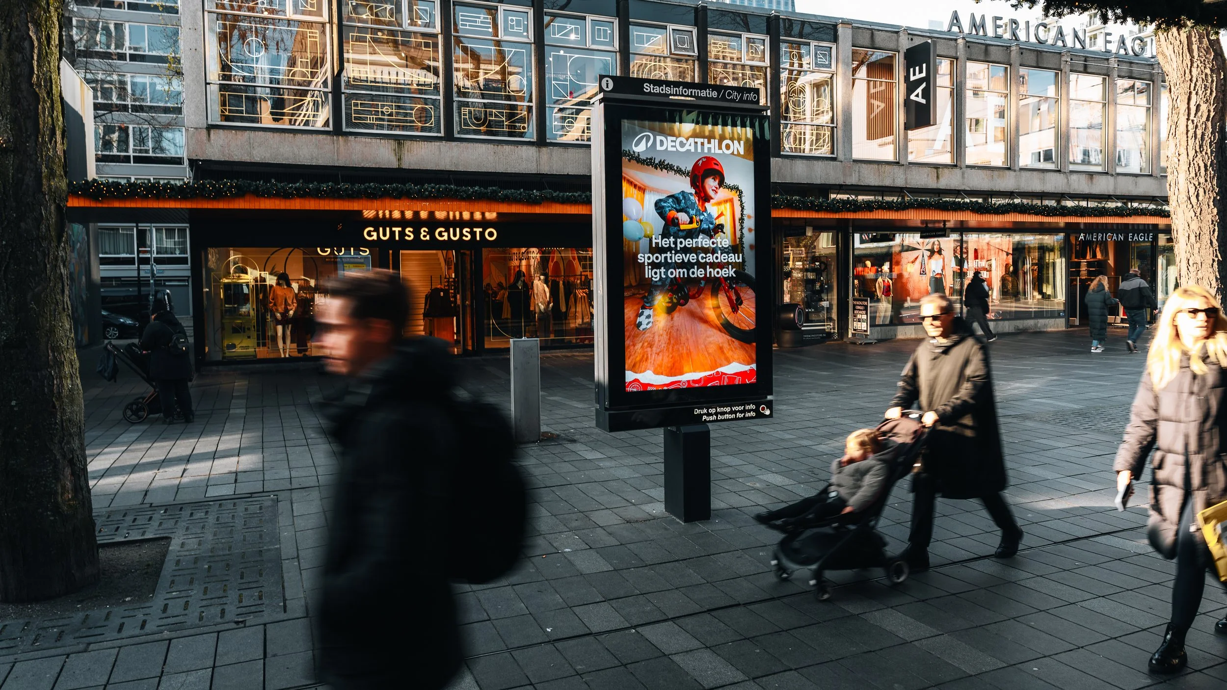 Een stadsplein met voetgangers, winkels en een digitale reclamezuil, waarop een kind op een fiets te zien is met de tekst 'Het perfecte sportieve cadeau ligt om de hoek'.
