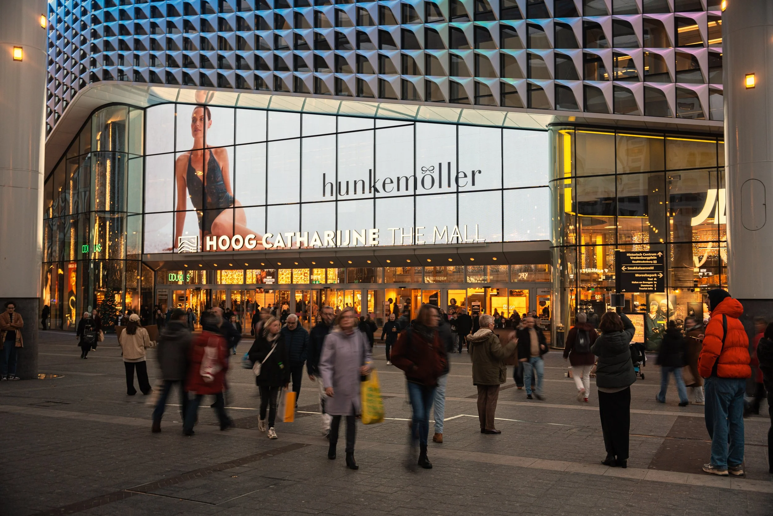 Een drukke winkelcentrum ingang met een groot digitale display en mensen die binnen en buiten lopen, met winkels en verlichte entree.