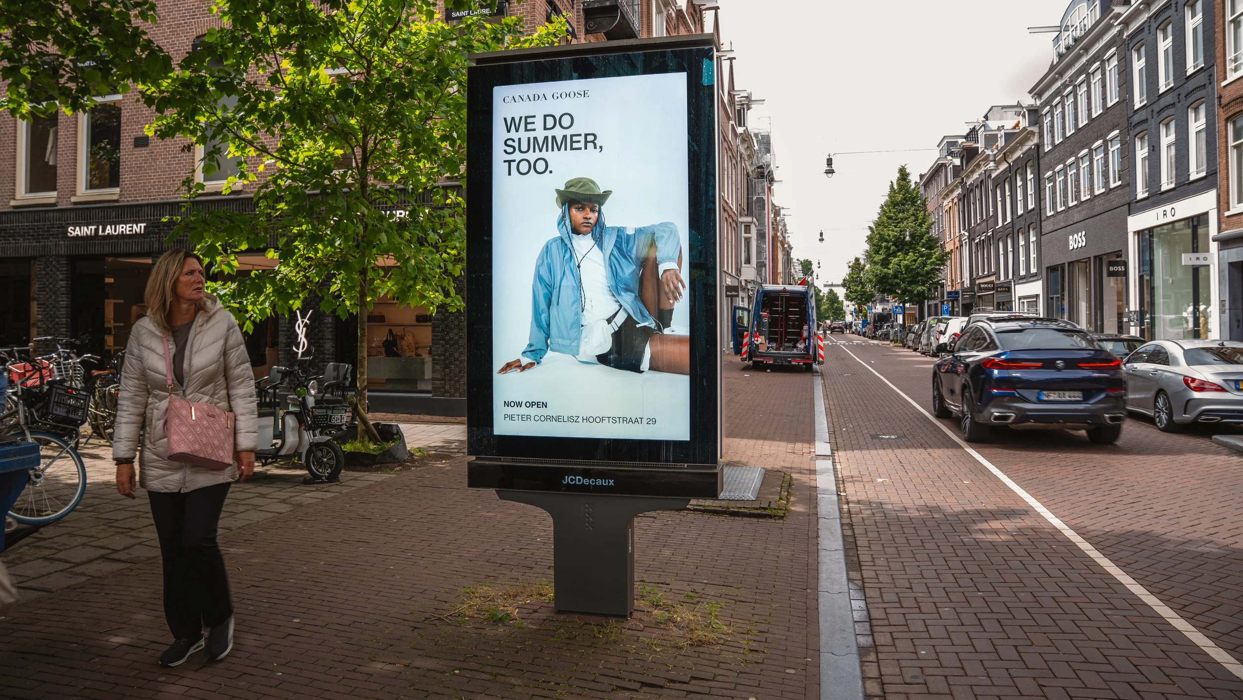 Straatbeeld met een digitale billboard die een modereclame toont voor een kledingwinkel, en een vrouw die wandelt met een roze tas, omgeven door winkels en geparkeerde auto's.