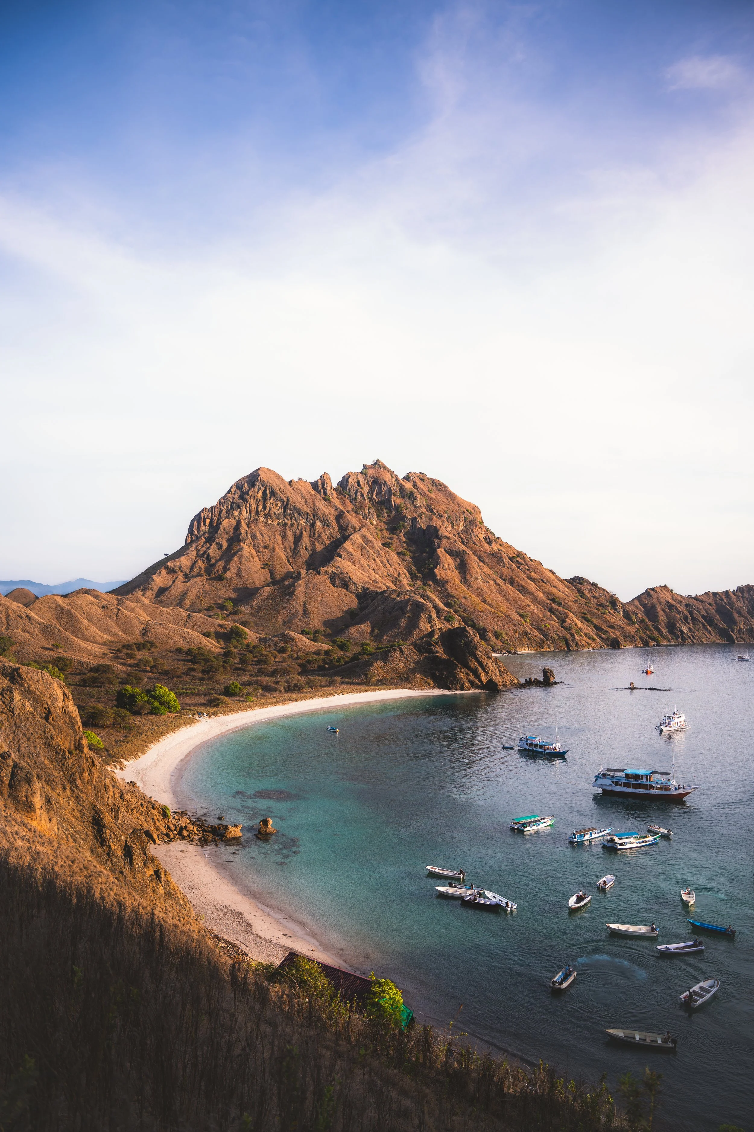 Een rustige baai met een zandstrand, omringd door bergen met droge vegetatie, en boten in het water.