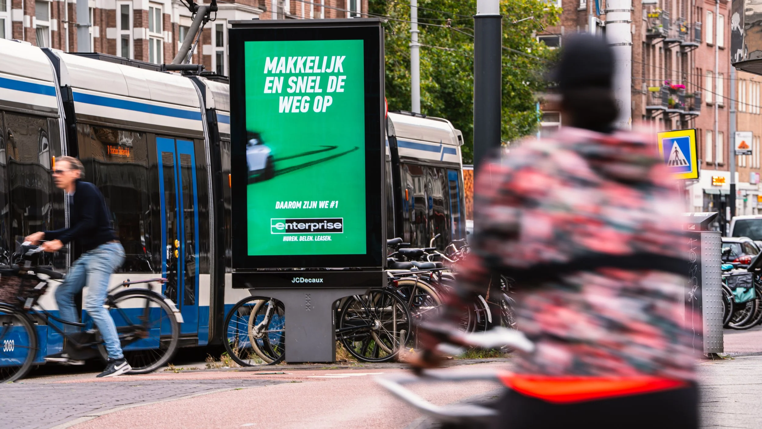 Een stadsbeeld met een digitale verkeersbord, fietsenrekken met fietsen, een tram, en een persoon die op een elektrische fiets rijdt. In de achtergrond zijn woongebouwen en een voetgangersoversteekplaats zichtbaar.