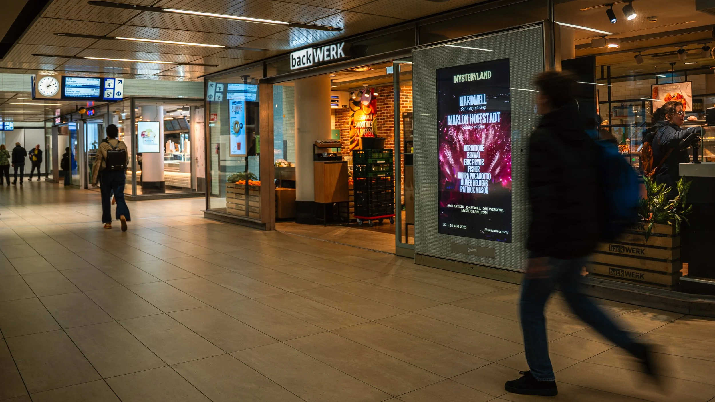 Binnenkomst in een treinstation met digitale informatieborden, winkels en mensen die lopen.