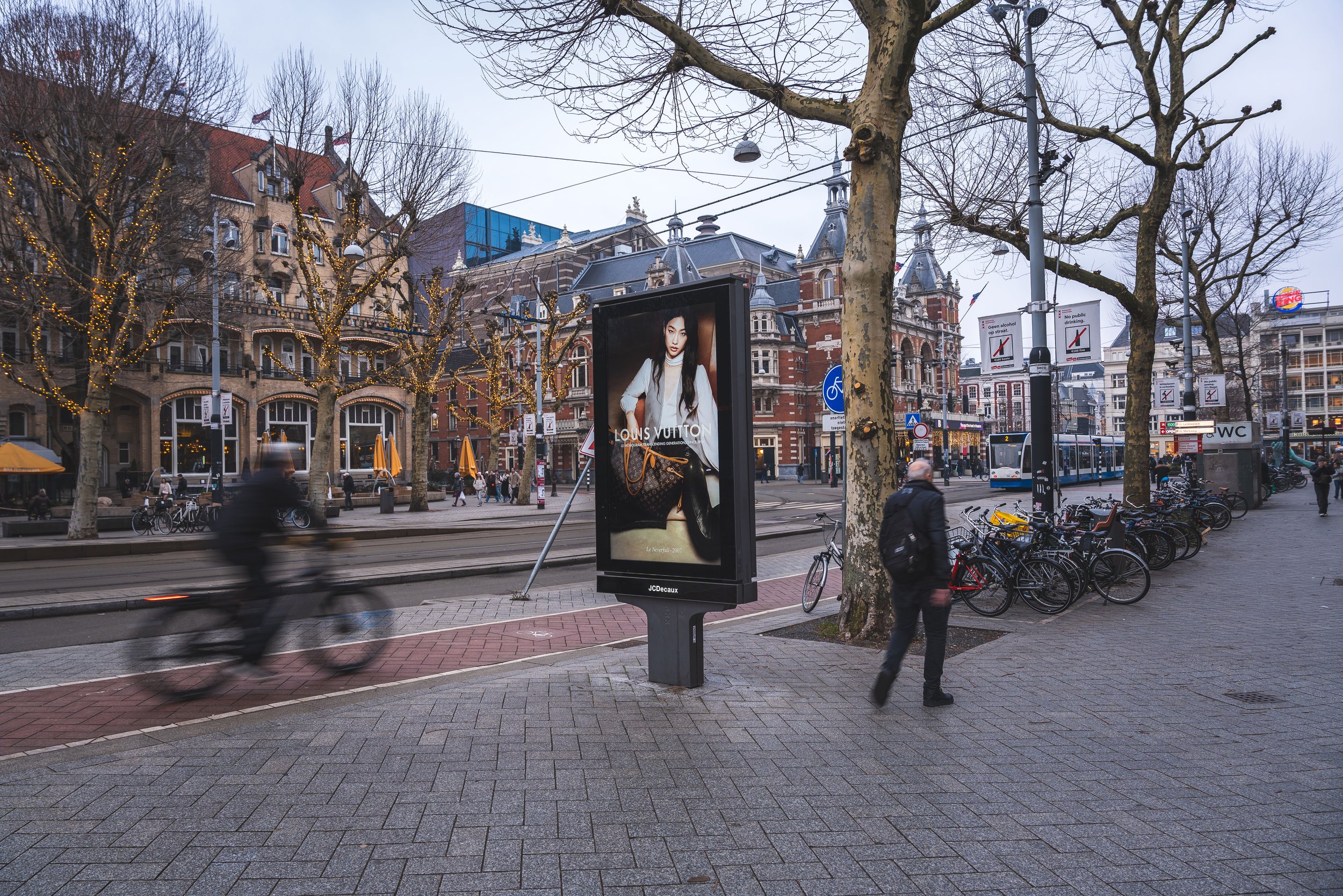 Een stadsplein met straatverlichting, bomen zonder bladeren, fietsen geparkeerd langs de stoep, voetgangers en een trambus in de achtergrond. Een digitaal billboard toont een advertisement van Louis Vuitton.
