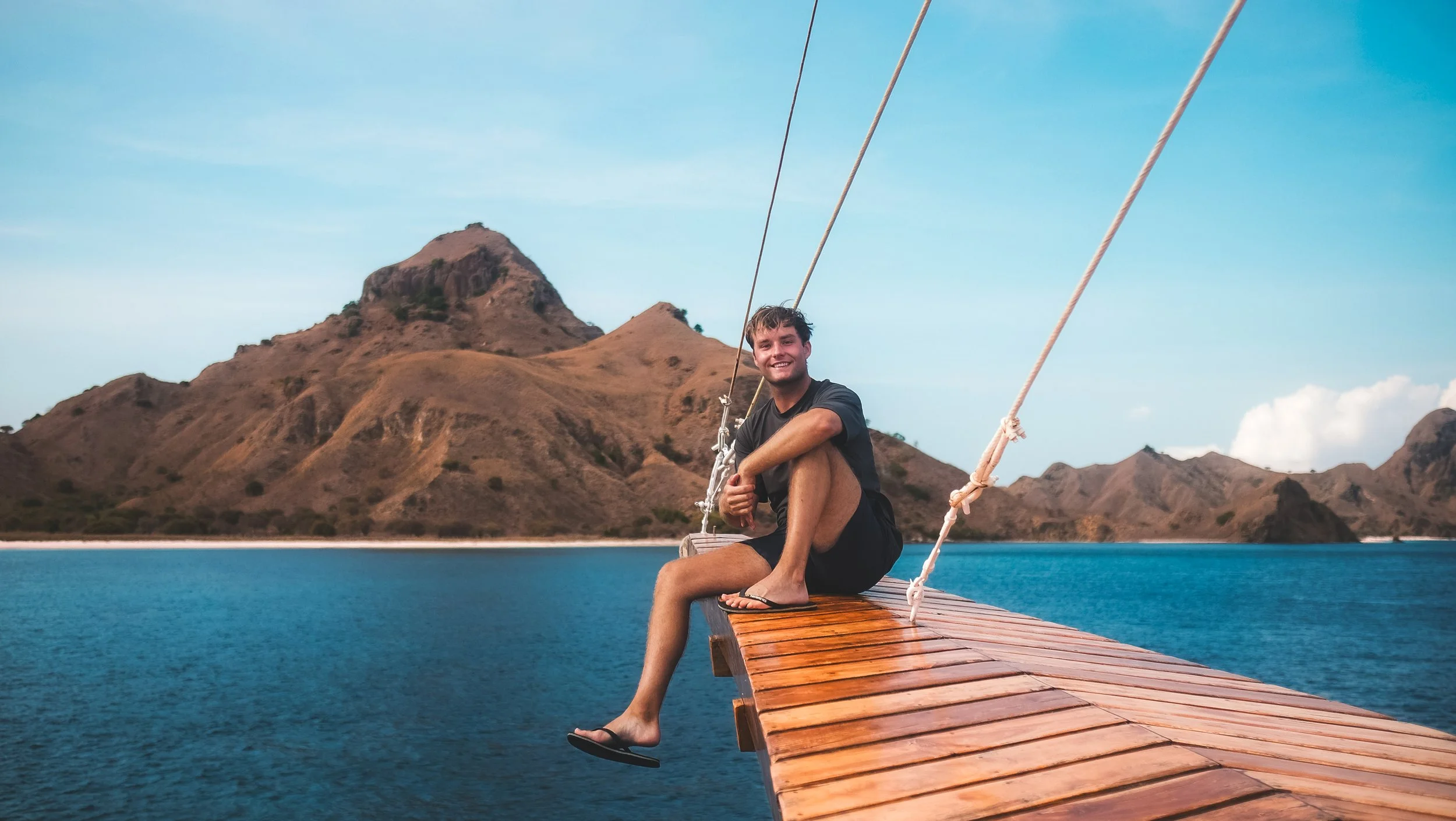 Een man zit ontspannend op een houten pier die uit een meer uitsteekt, met bergen op de achtergrond en een heldere blauwe hemel.