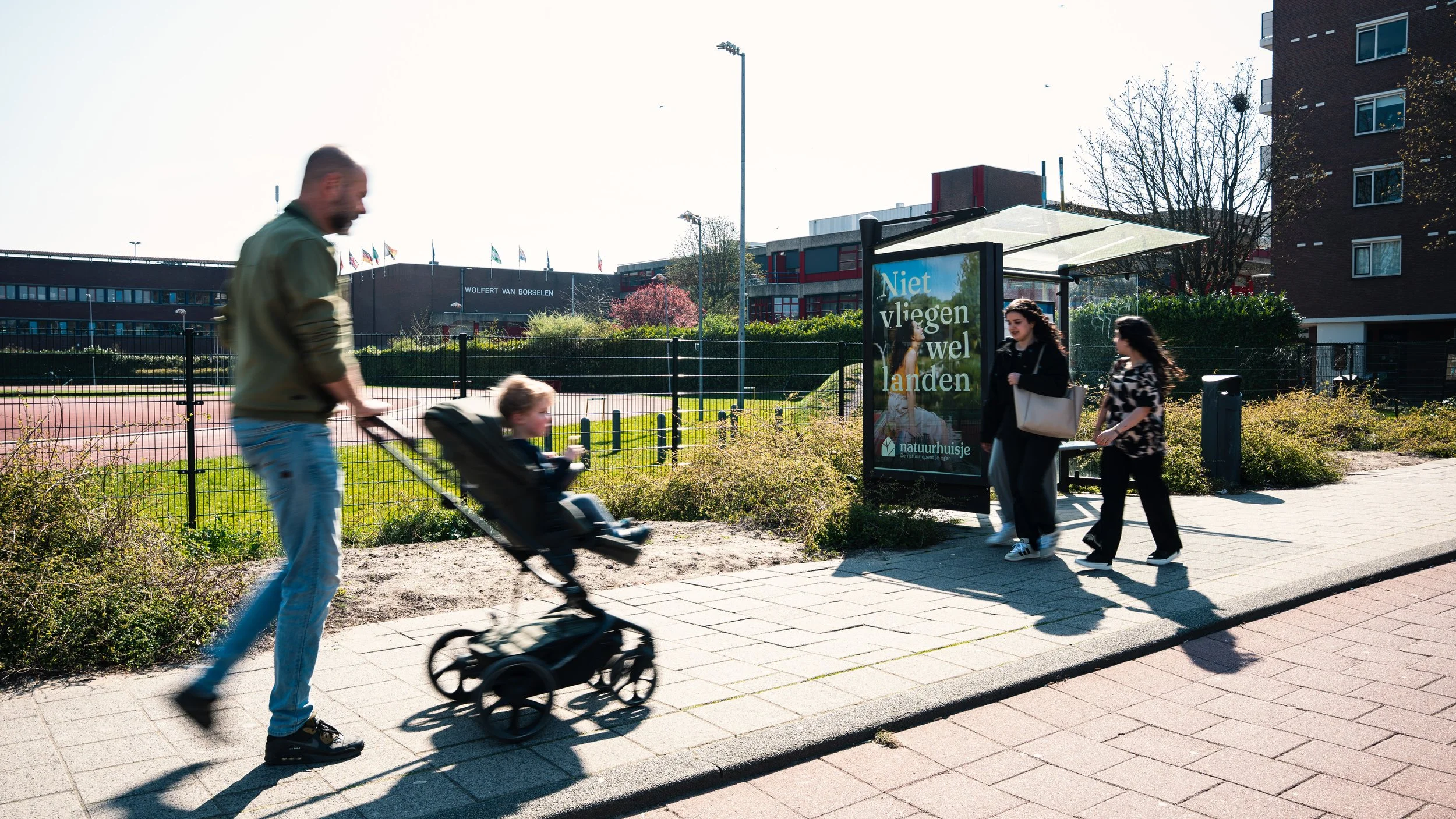 Mensen wachten bij een bushalte, inclusief een man die een kind in een kinderwagen duwt en twee vrouwen die de halte naderen. Op de bushalte staat een reclamebord met de tekst 'Niet vliegen wel landen'.