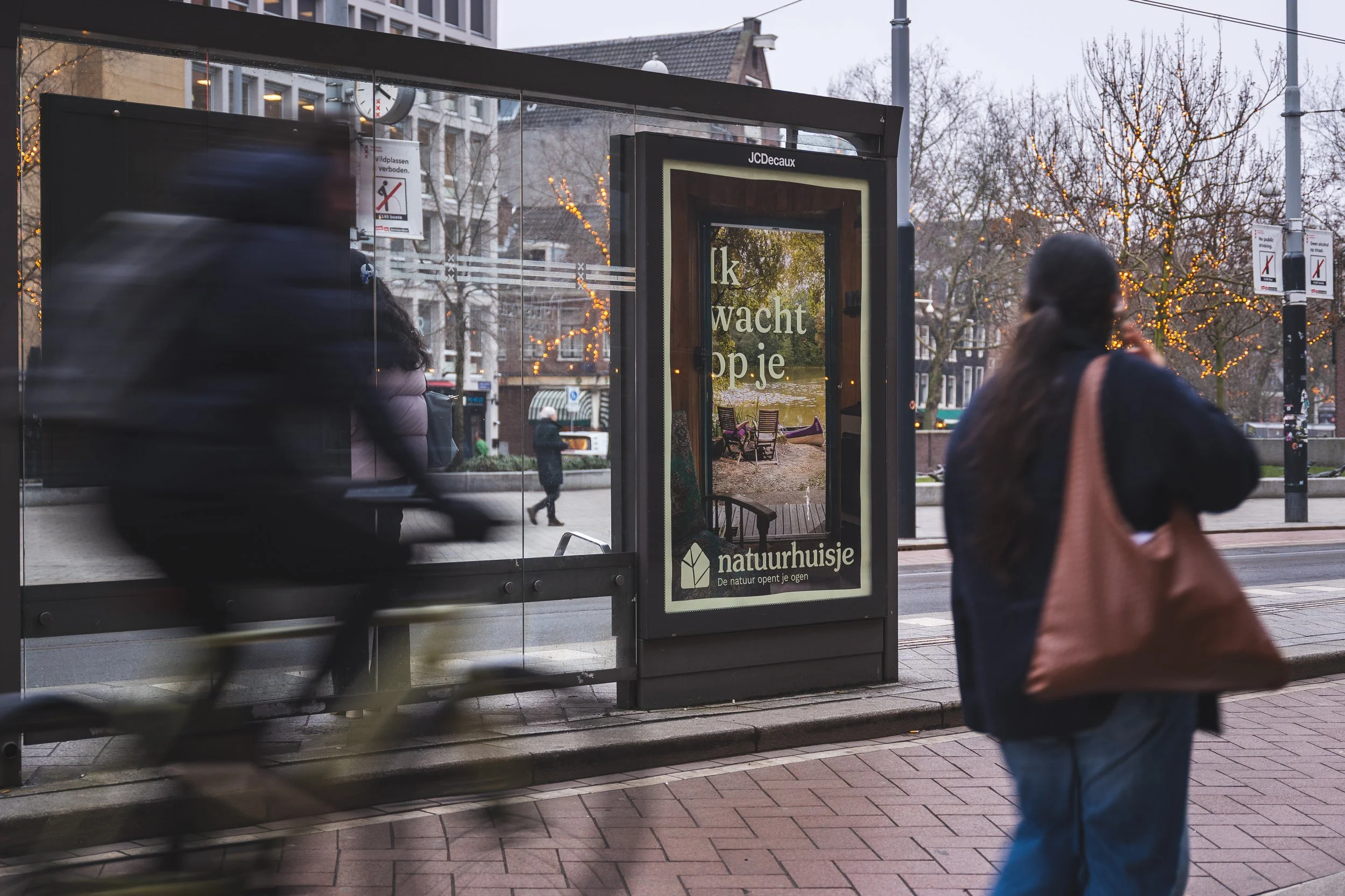 Een bushalte in de stad met een digitaal bord dat zegt: 'Ik wacht op je.' Op de achtergrond is een park met een bank en bomen met kerstverlichting. Een fietser rijdt langs de halte en een persoon loopt met een tas.