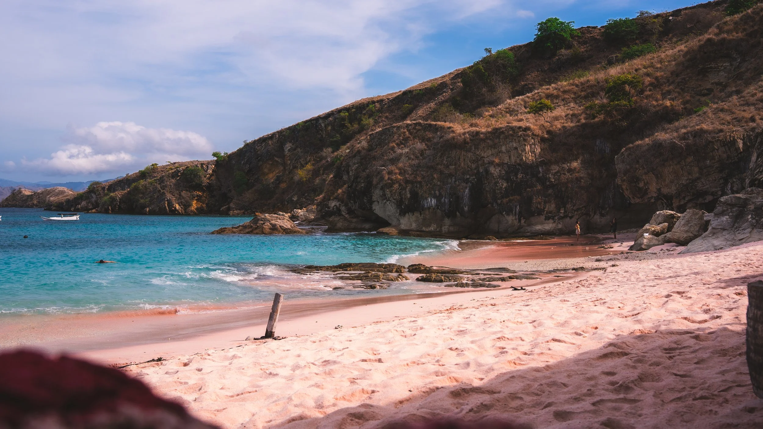 Een rustig strand met witte zand, groene heuvels en blauwe zee, met twee personen die wandelen en een kleine boot op het water.