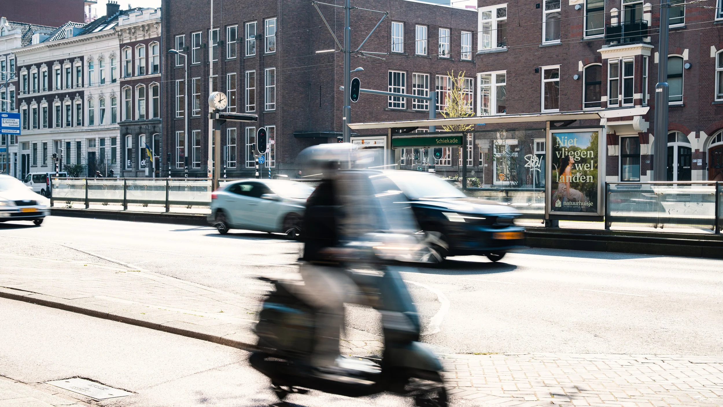 Een straat in de stad met een tramhalte en een bushokje. Verschillende auto's en een scooter rijden door de straat. De gebouwen op de achtergrond zijn oud en hebben grote ramen.
