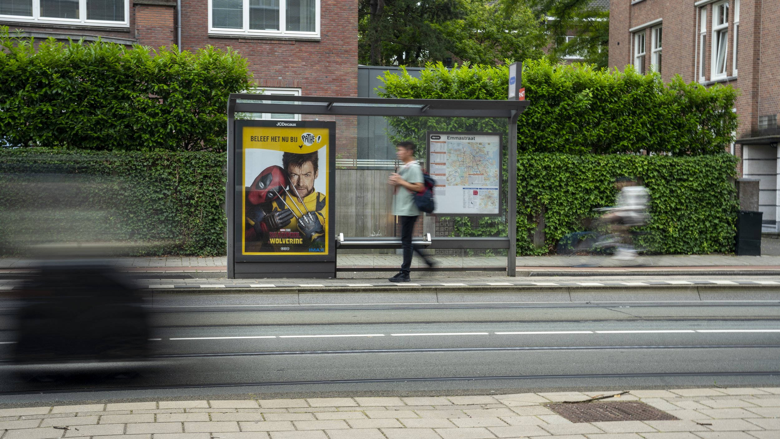Een bushalte met een poster van Deadpool en een plattegrond van de metro. Een man met rugzak staat te wachten, terwijl auto's en een motorrijder snel voorbijrijden.