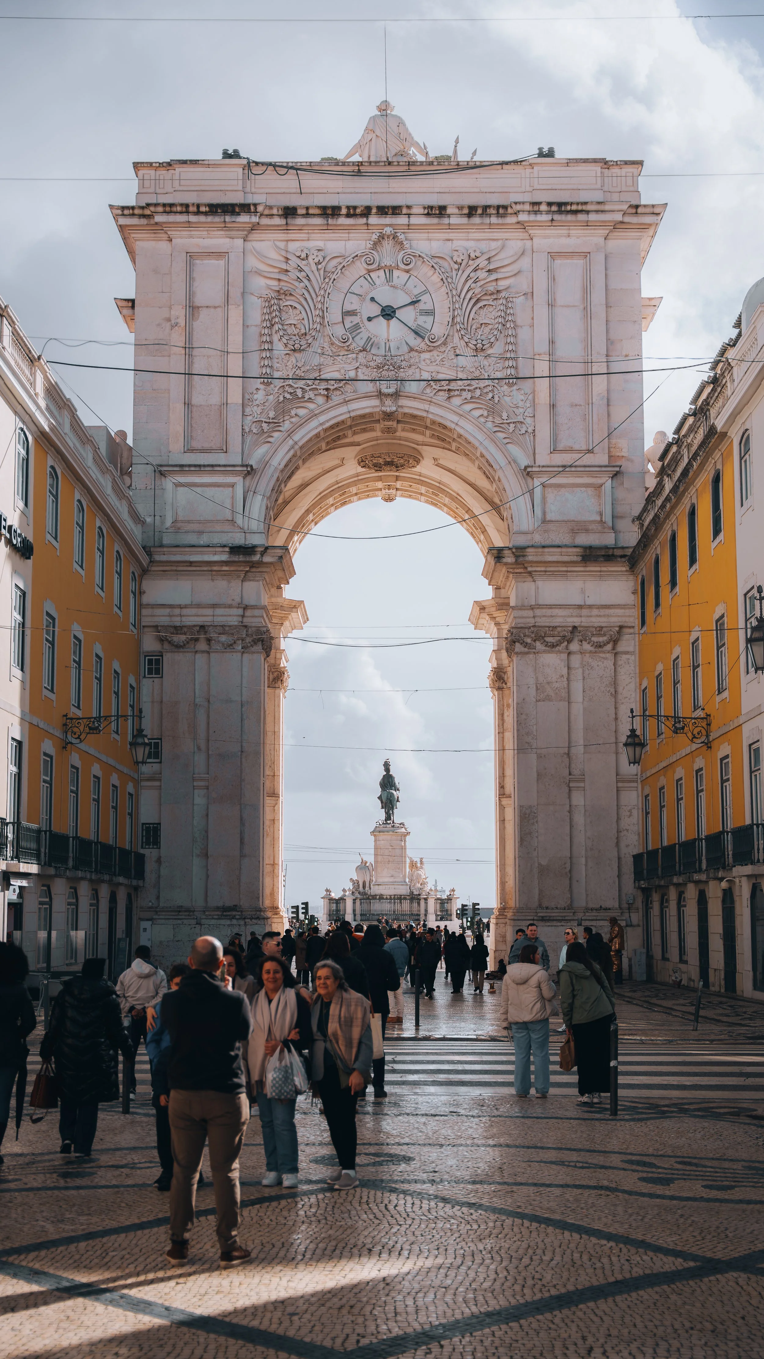 Arch met een klok en beeldhouwwerk, bezoekers op straat, zicht op standbeeld op de achtergrond in Lissabon.