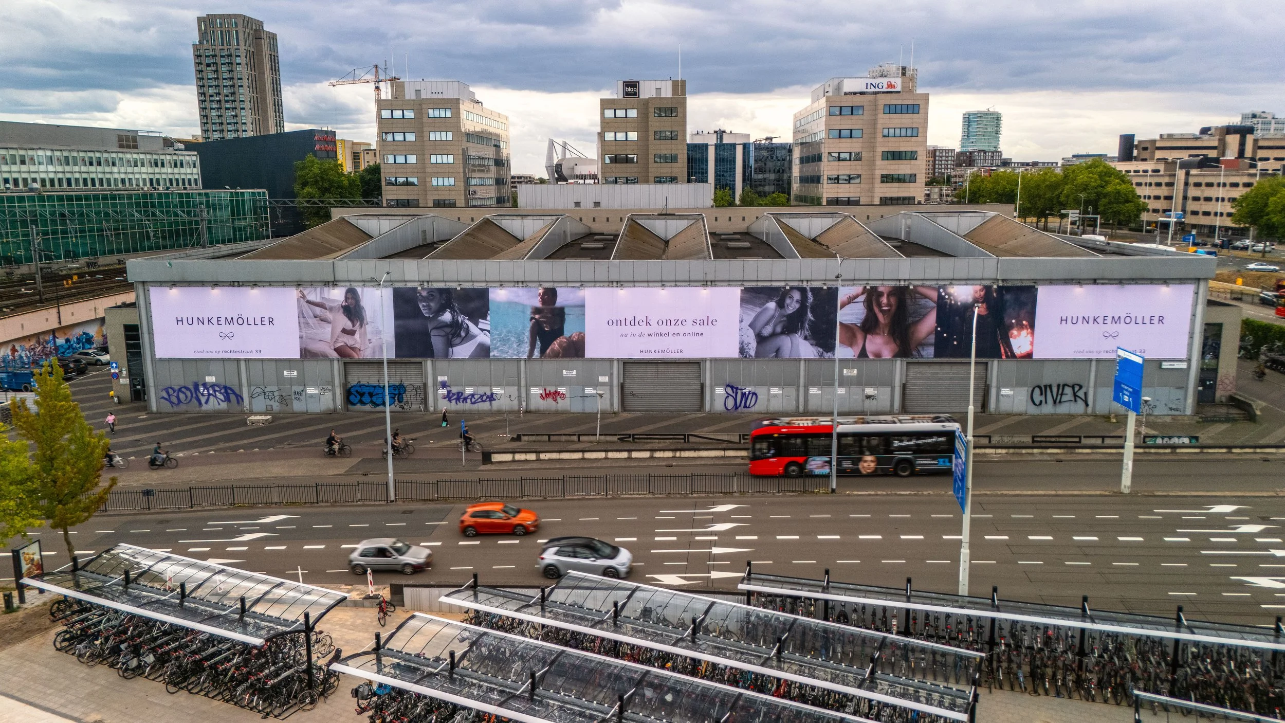 Stedelijk gebouw met advertenties voor Hunkemöller en diverse graffiti op de onderkant van de gevel, een weg met vier rijstroken en auto’s, een bus en fietsers, en een fietsstalling op de voorgrond onder een bomenrij, in een stedelijke omgeving met h