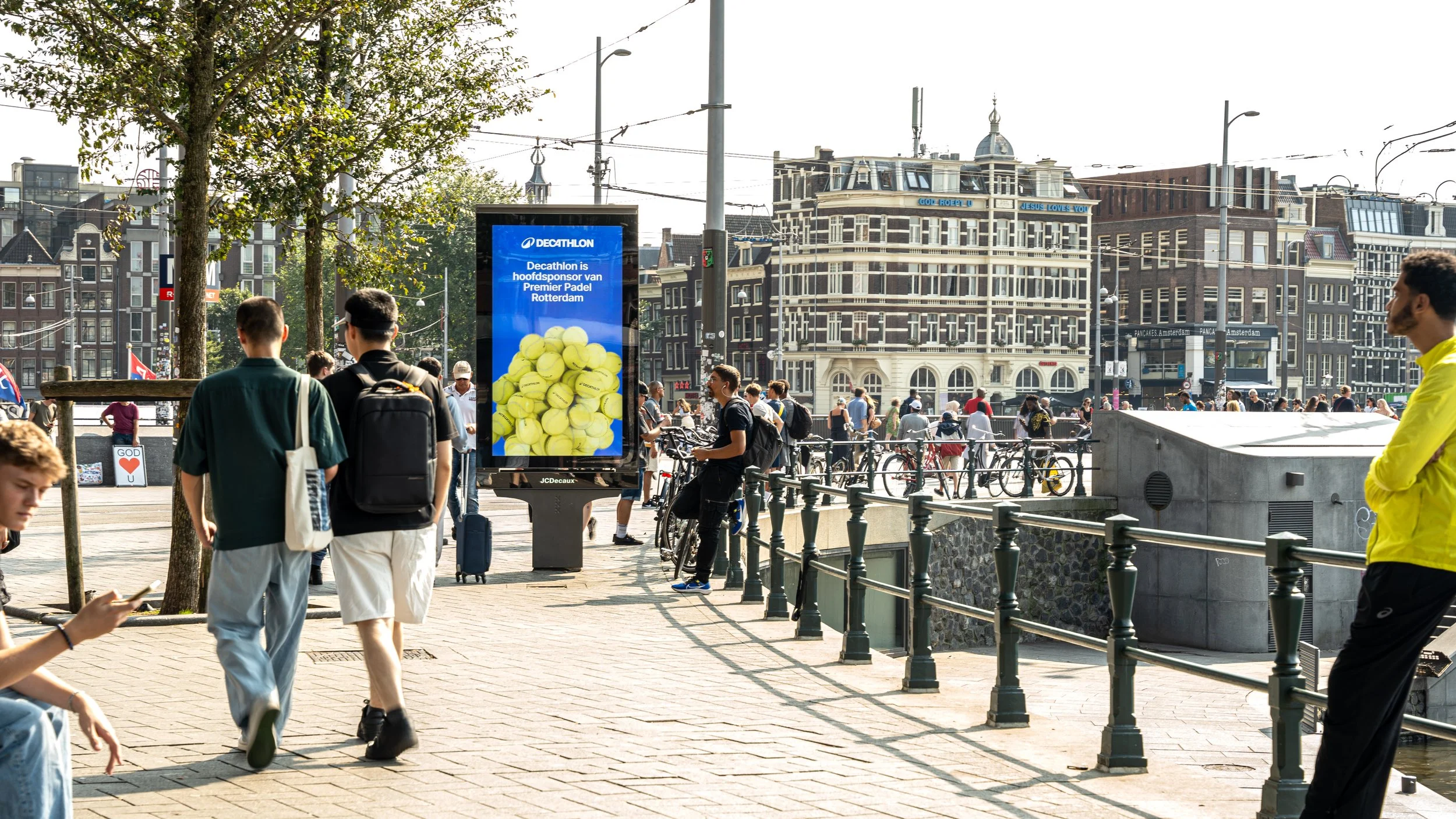 Straatbeeld in Amsterdam met mensen, een digitale poster met tennisballen, oude gebouwen op de achtergrond en een heldere hemel.
