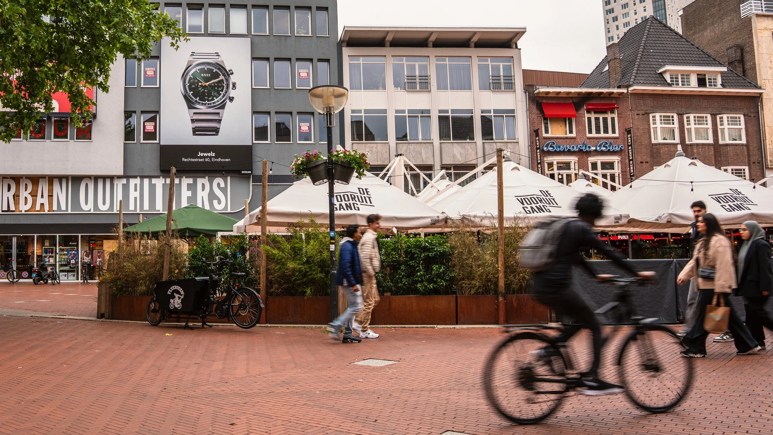 Stadsplein met wandelaars en fietser, terrassen met parasols, moderne gebouwen en een groot horlogeadvertentie op een gebouw.