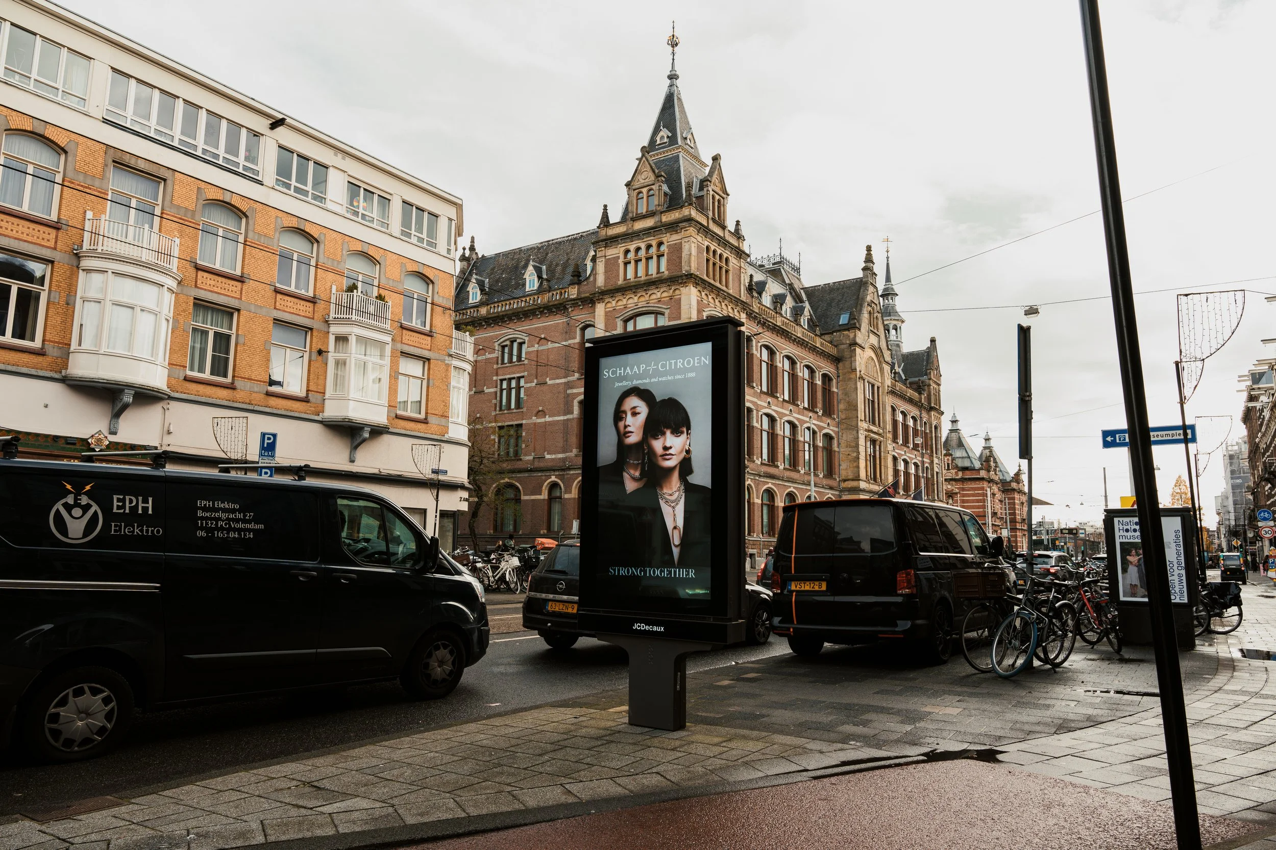 Straatbeeld met geparkeerde auto's, fietsen, reclamebord en historische gebouwen in een Europees stadscentrum.