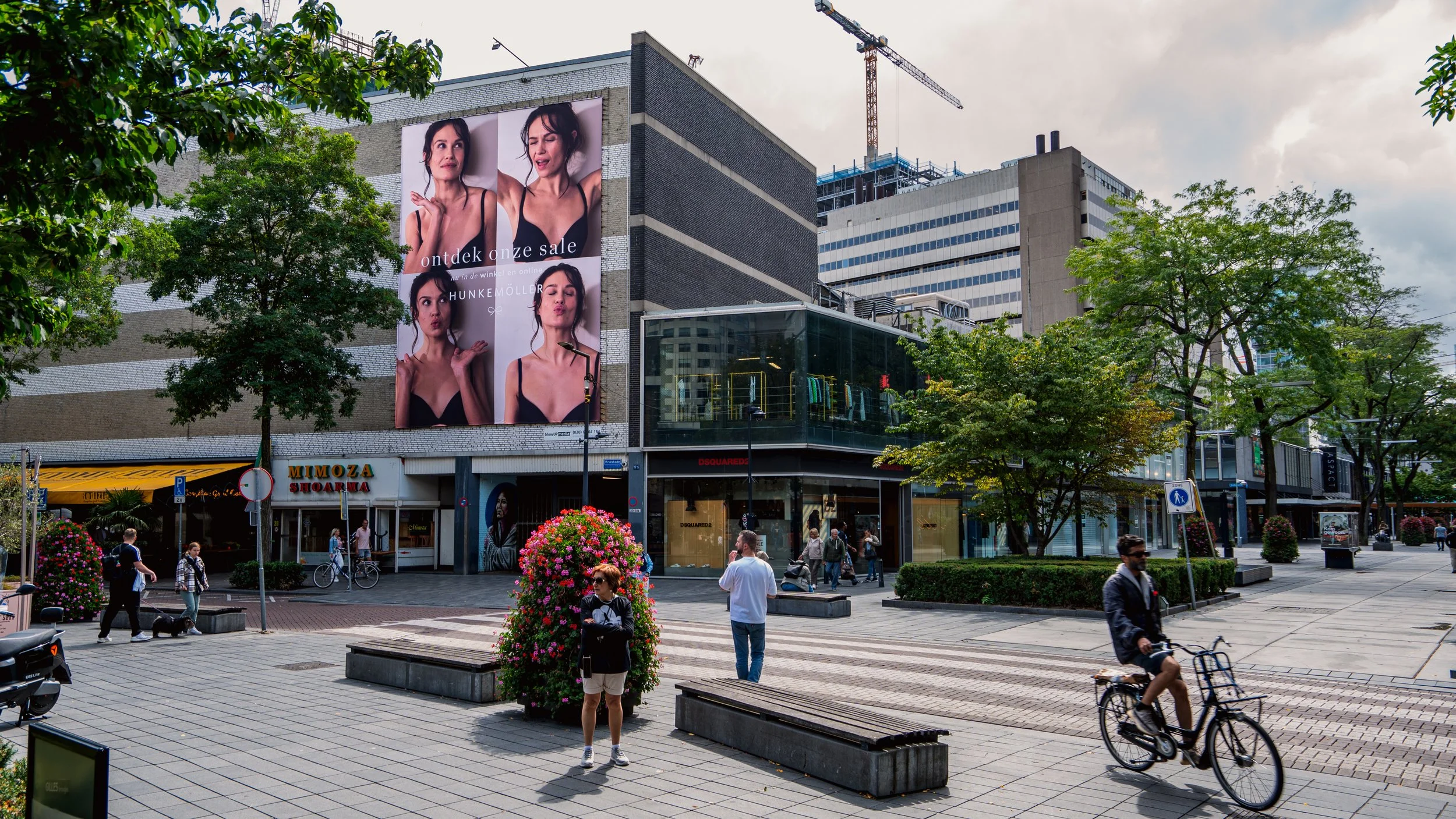 Een stadsplein met winkelpanden, groene bomen, mensen die lopen en fietsen, een grote advertentiebillboard, en bloemen in bloembedden.
