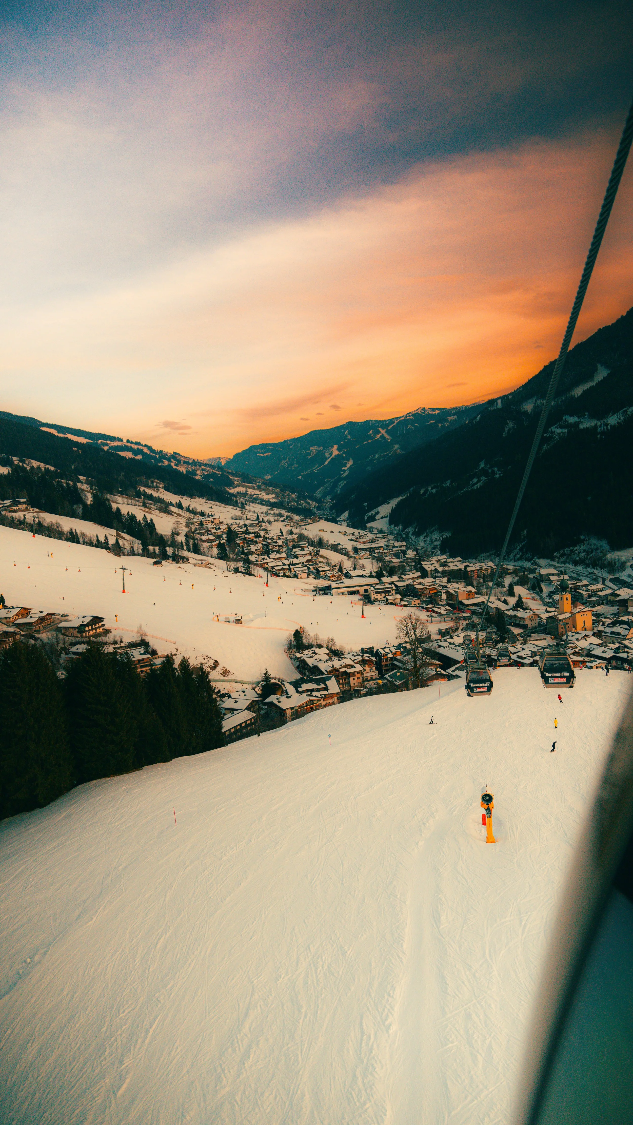 Skigondelbaan die omhoog gaat met uitzicht op besneeuwde bergen, een dorp en een kleurrijke zonsondergang in de achtergrond.