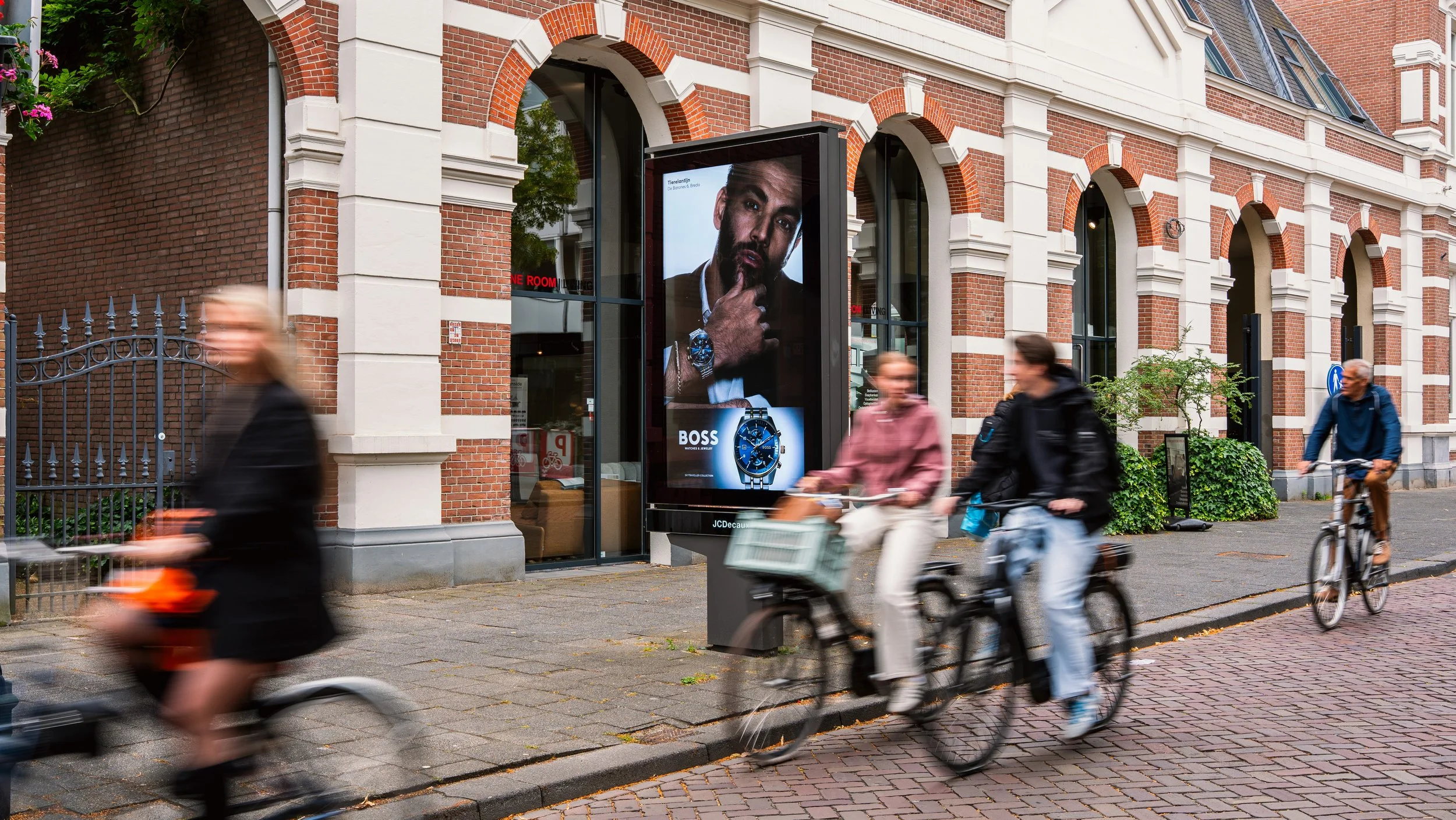 Twee mannen en een vrouw fietsen langs een oude bakstenen gebouw met advertentieborden, een roze jas en een zwarte jas, een man met een blauwe jas en een fiets.