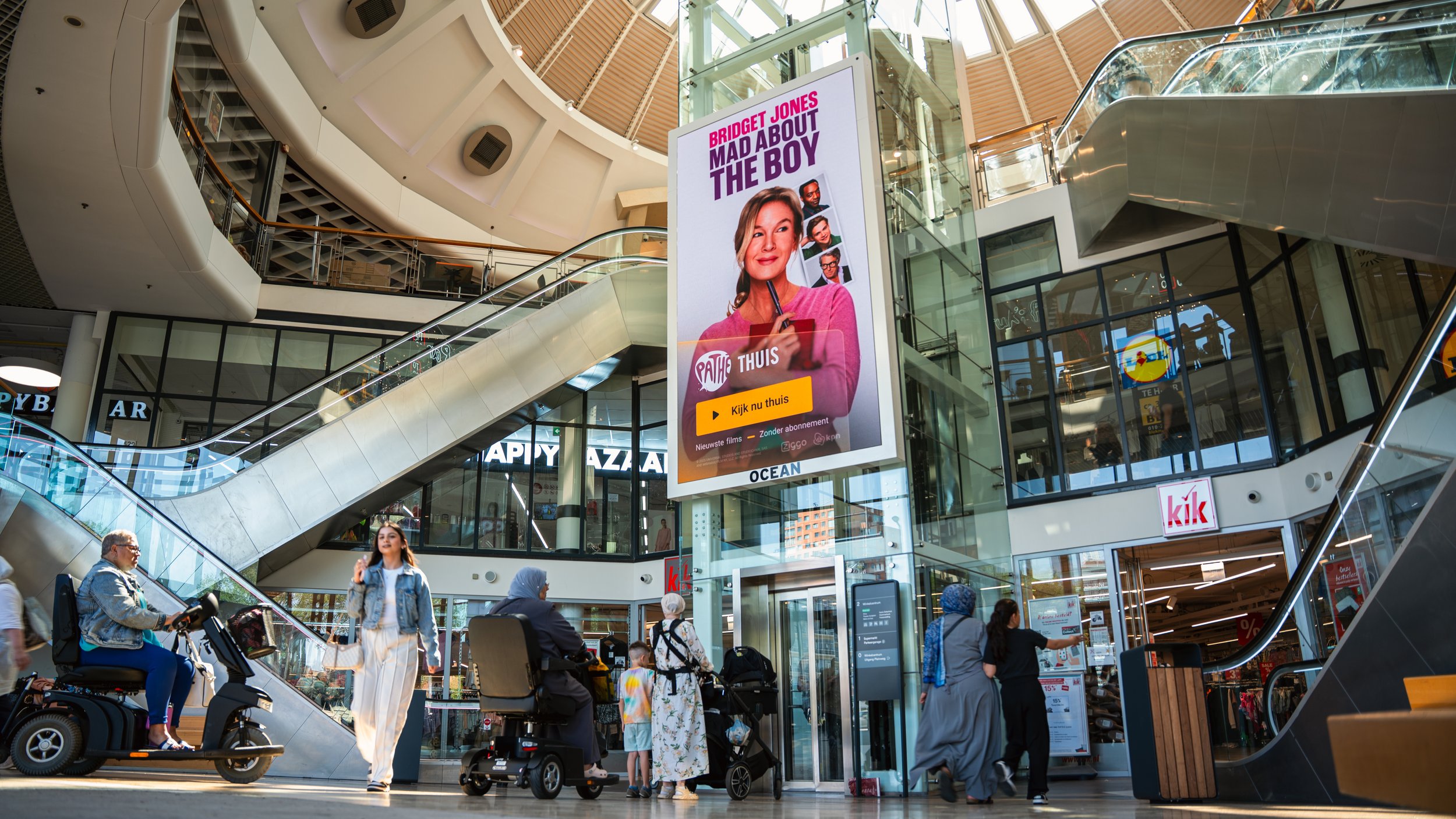 Interieur van een winkelcentrum met grote digitale reclameborden, mensen die rondlopen, sommige met rollators, en een persoon op een scooter.