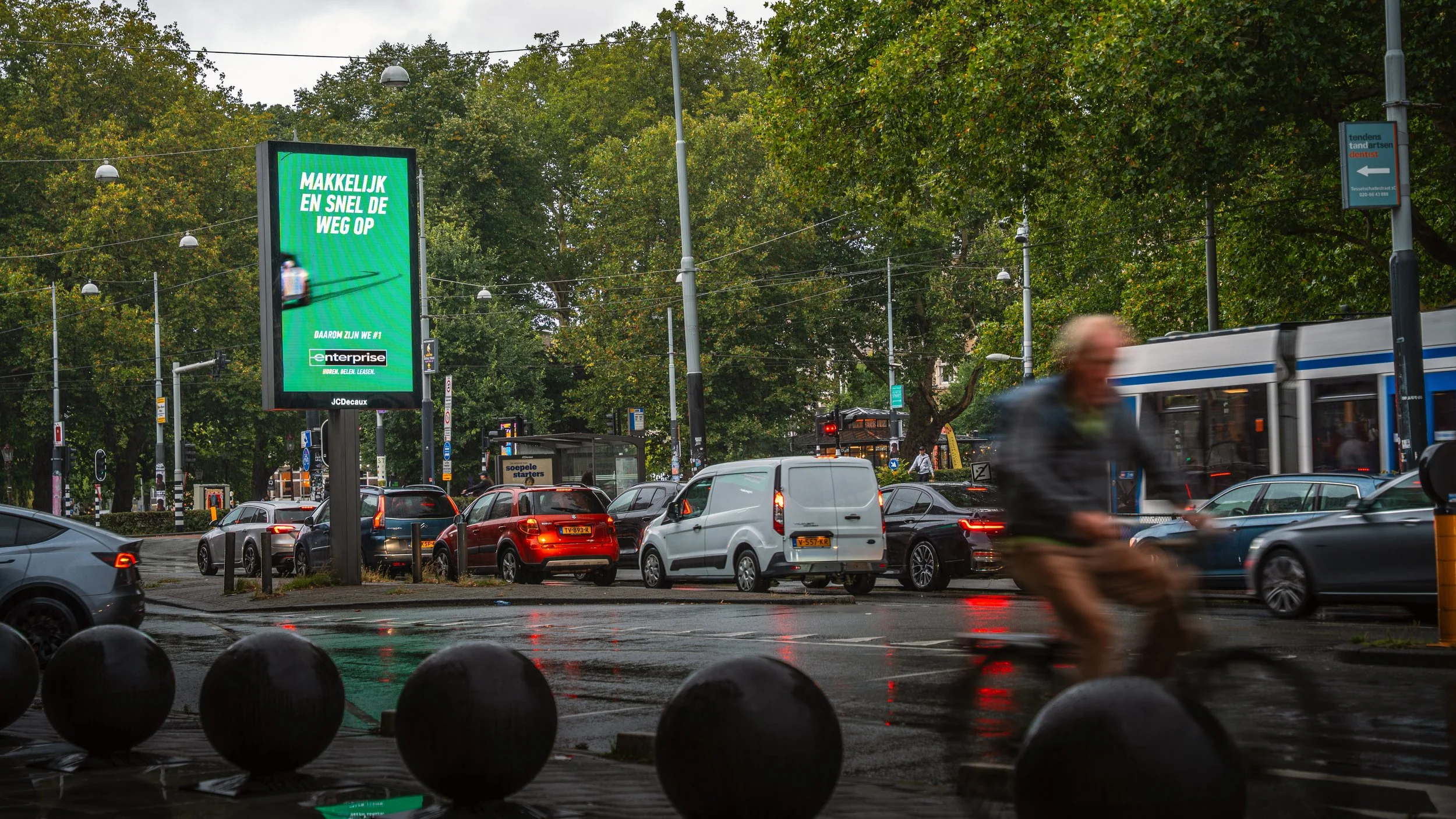 Een drukke straat met auto's, een tram en een fietser, terwijl het regent, met groene bomen op de achtergrond en een digitale reclamebord dat in het Nederlands reclame maakt.