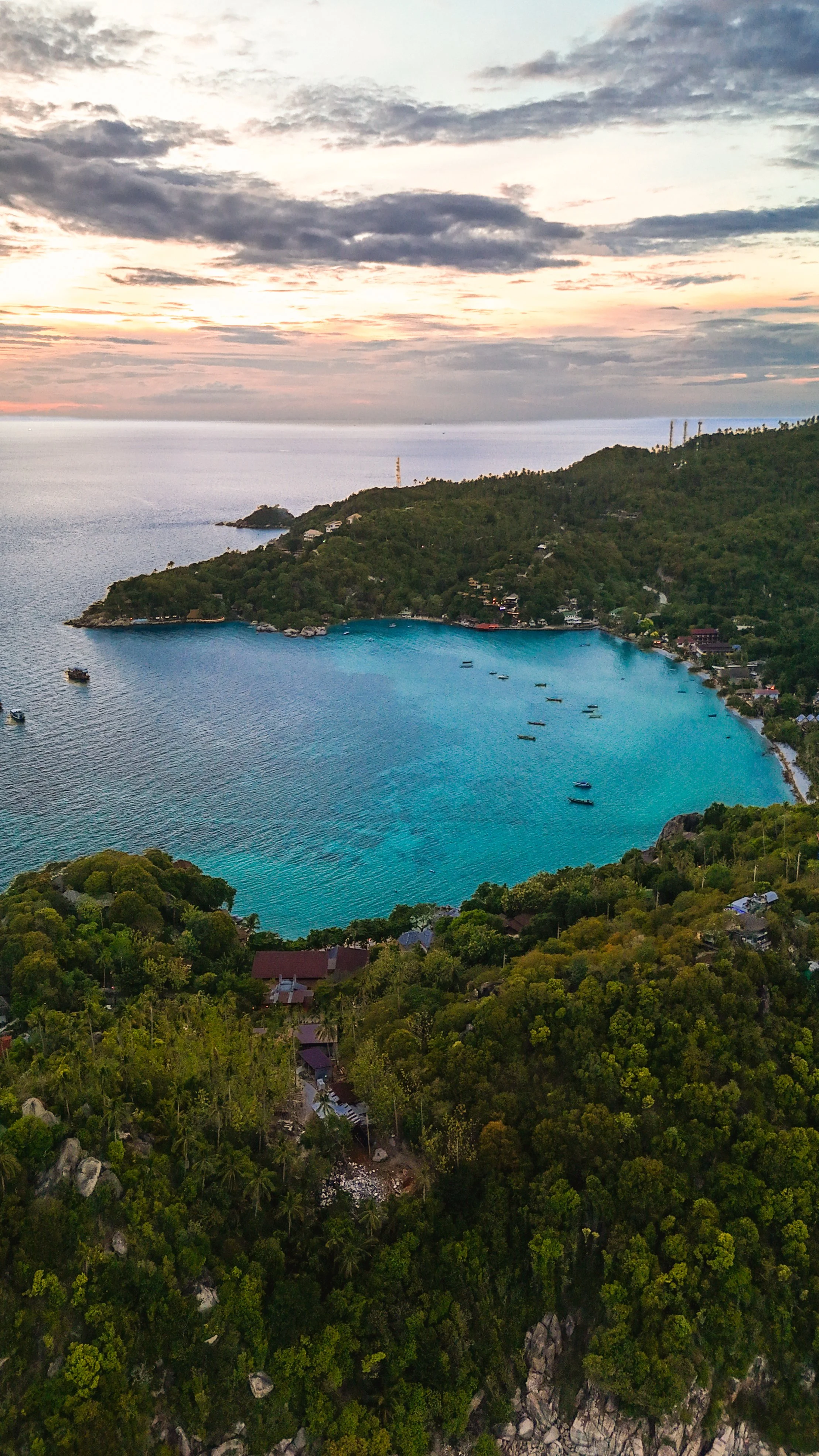Luchtfoto van een baai met helderblauw water, omgeven door groene heuvels en bebouwde gebieden, onder een bewolkte hemel tijdens zonsondergang.