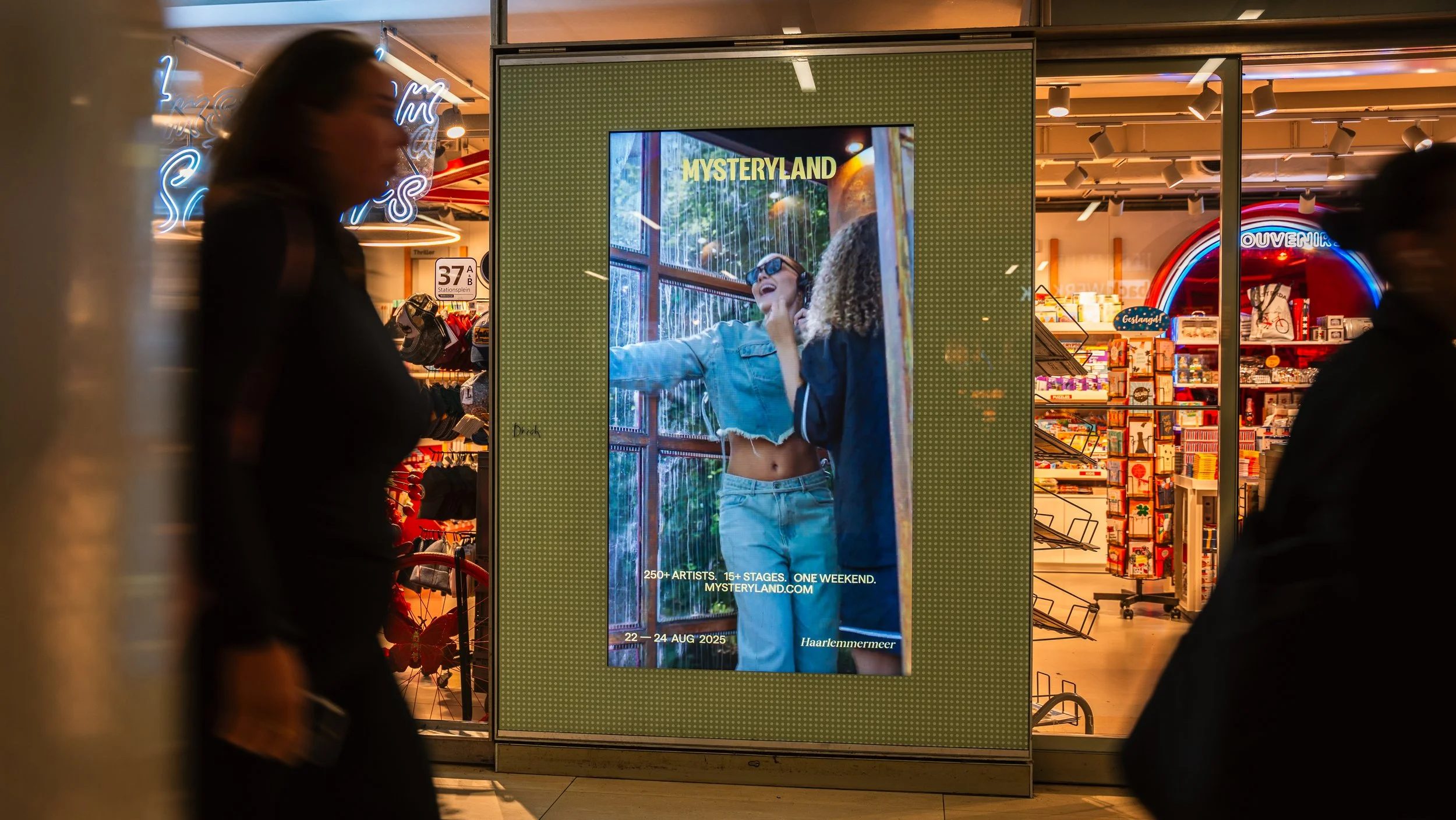 Elektronische display met een afbeelding van twee jonge vrouwen die lachen en plezier hebben, met tekst over Mythicaland muziekfestival, dat plaatsvindt van 22 tot 24 augustus 2025, in Haarlemmermermeer.