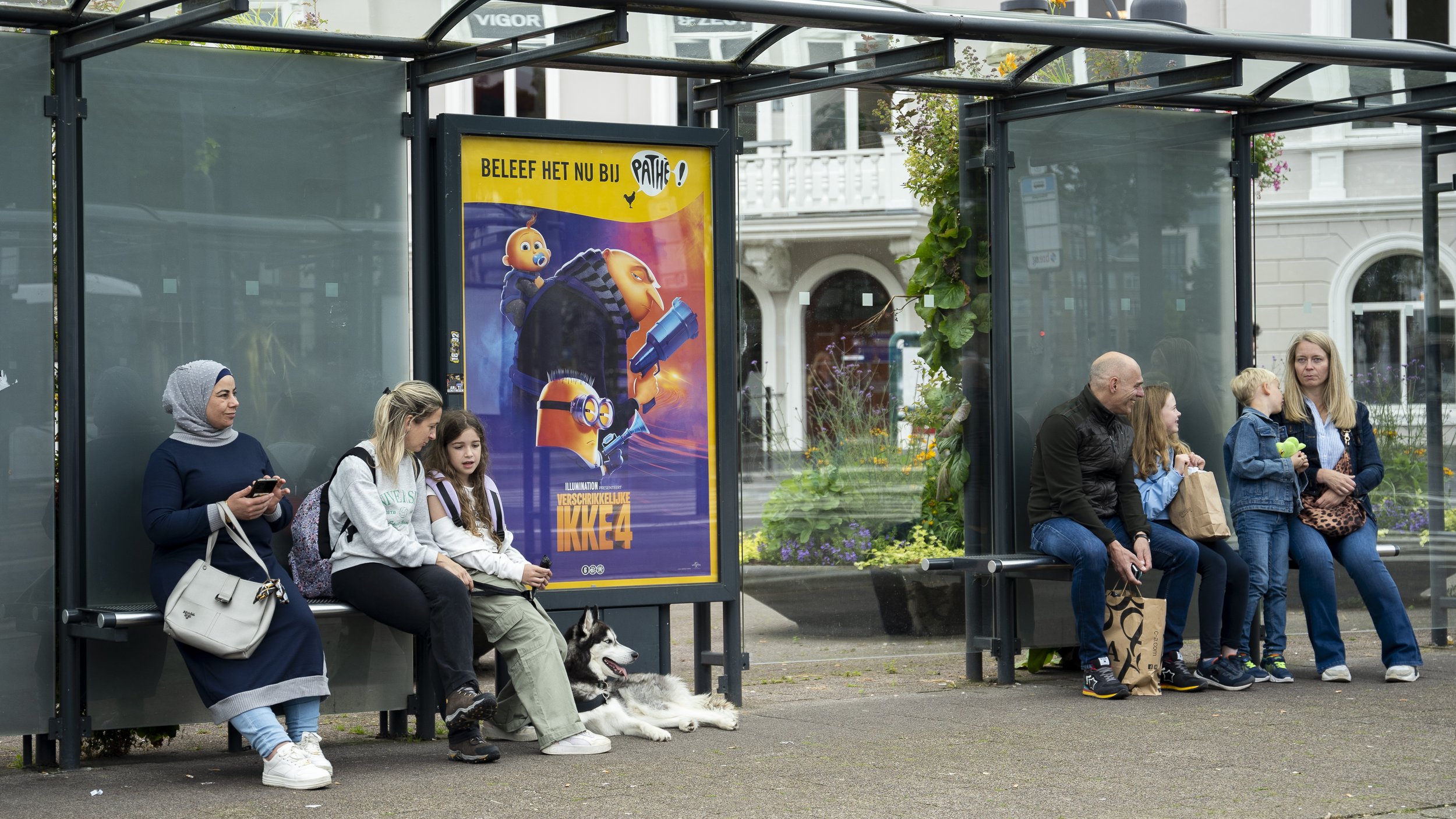 Twee groepen mensen wachten bij een bushalte, met een poster voor een film met cartoonpersonages erachter. Links zitten een vrouw met hoofddoek, twee jonge meisjes en een husky. Rechts zitten een man, een meisje, een jongen en een vrouw.