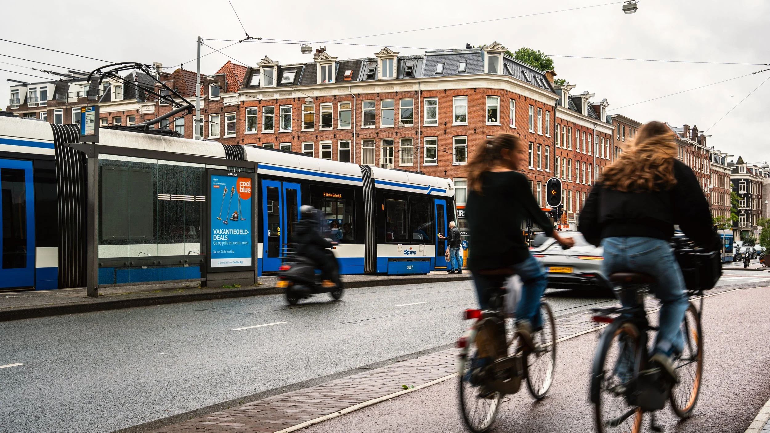 Twee mensen fietsen langs een tramhalte met een tram in een stedelijk gebied met oude gebouwen op de achtergrond, tijdens een bewolkte dag.