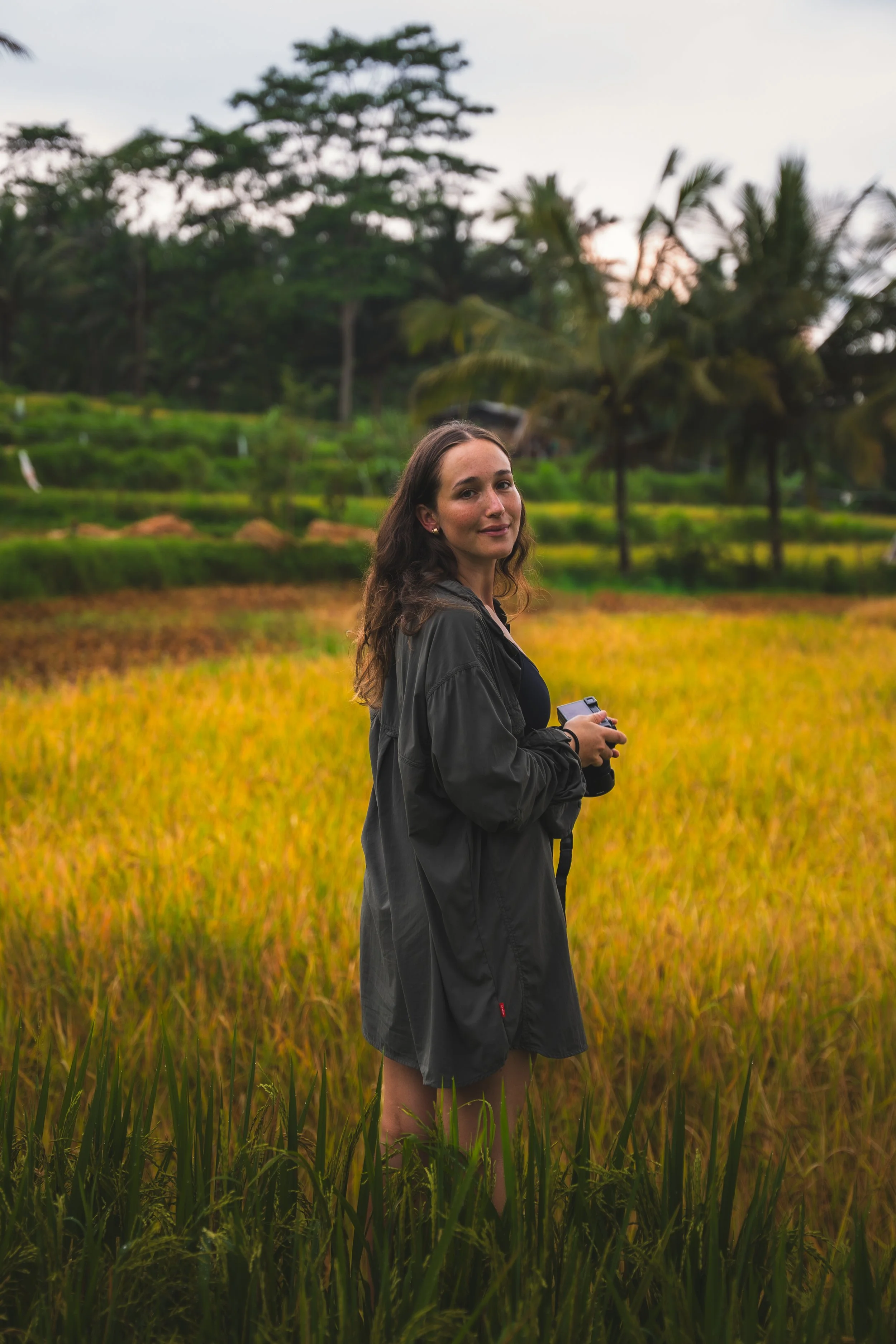 Een vrouw met bruin haar, gekleed in een donkergrijze jas, staat in een veld met rijstplanten, terwijl ze een camera vasthoudt. Op de achtergrond bevinden zich groene bomen en palmbomen onder een bewolkte hemel.