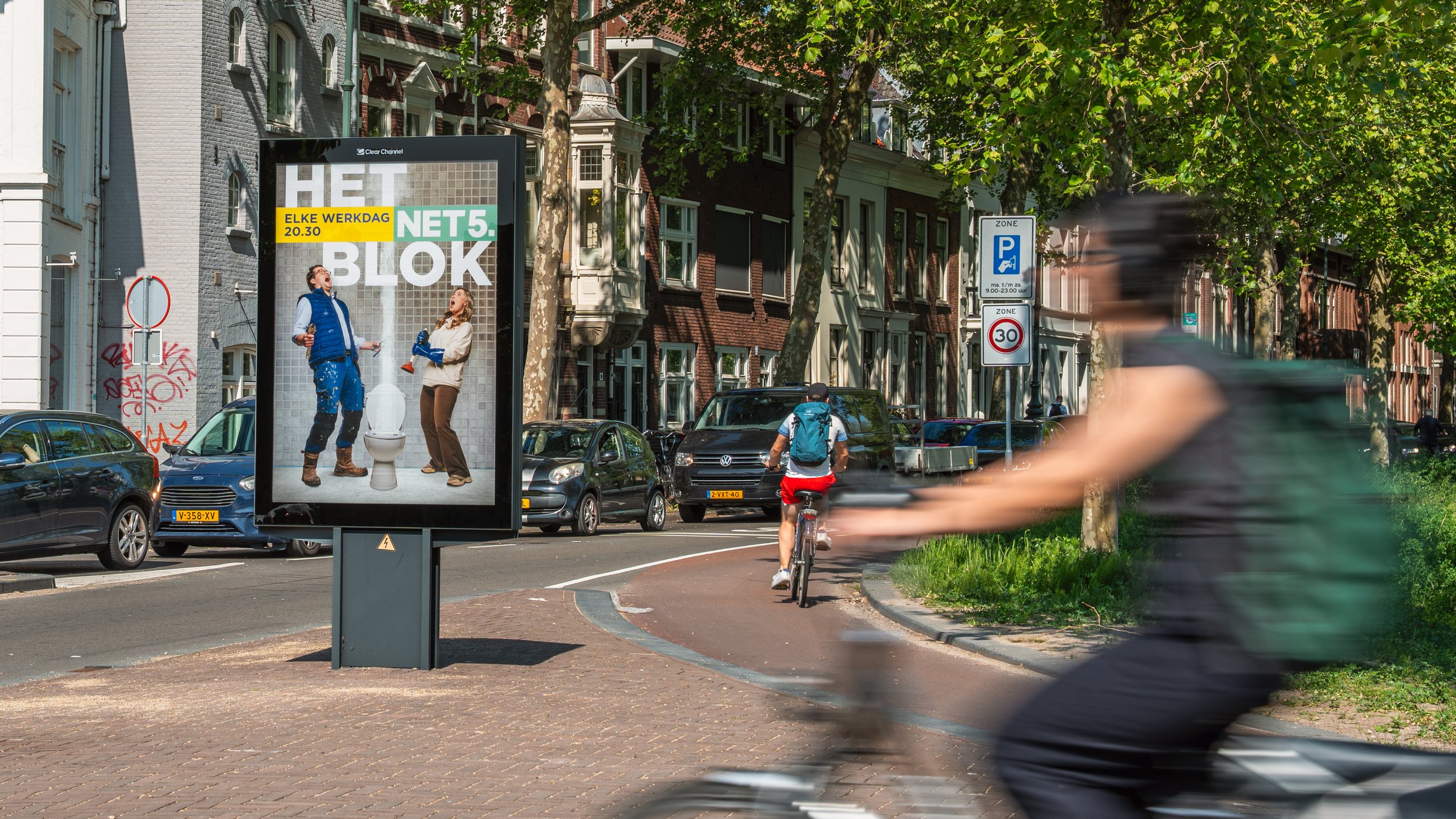 Verkeersplein met fietsers en auto's, groene bomen, gevels van huizen, en een digitale billboard met een advertentie.