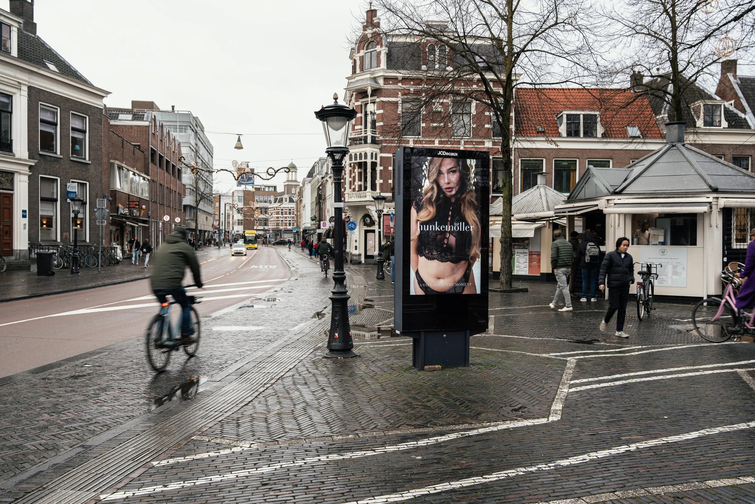Straat met fietsers, mensen wandelen, reclamebord met model, historische gebouwen, gemengd weer