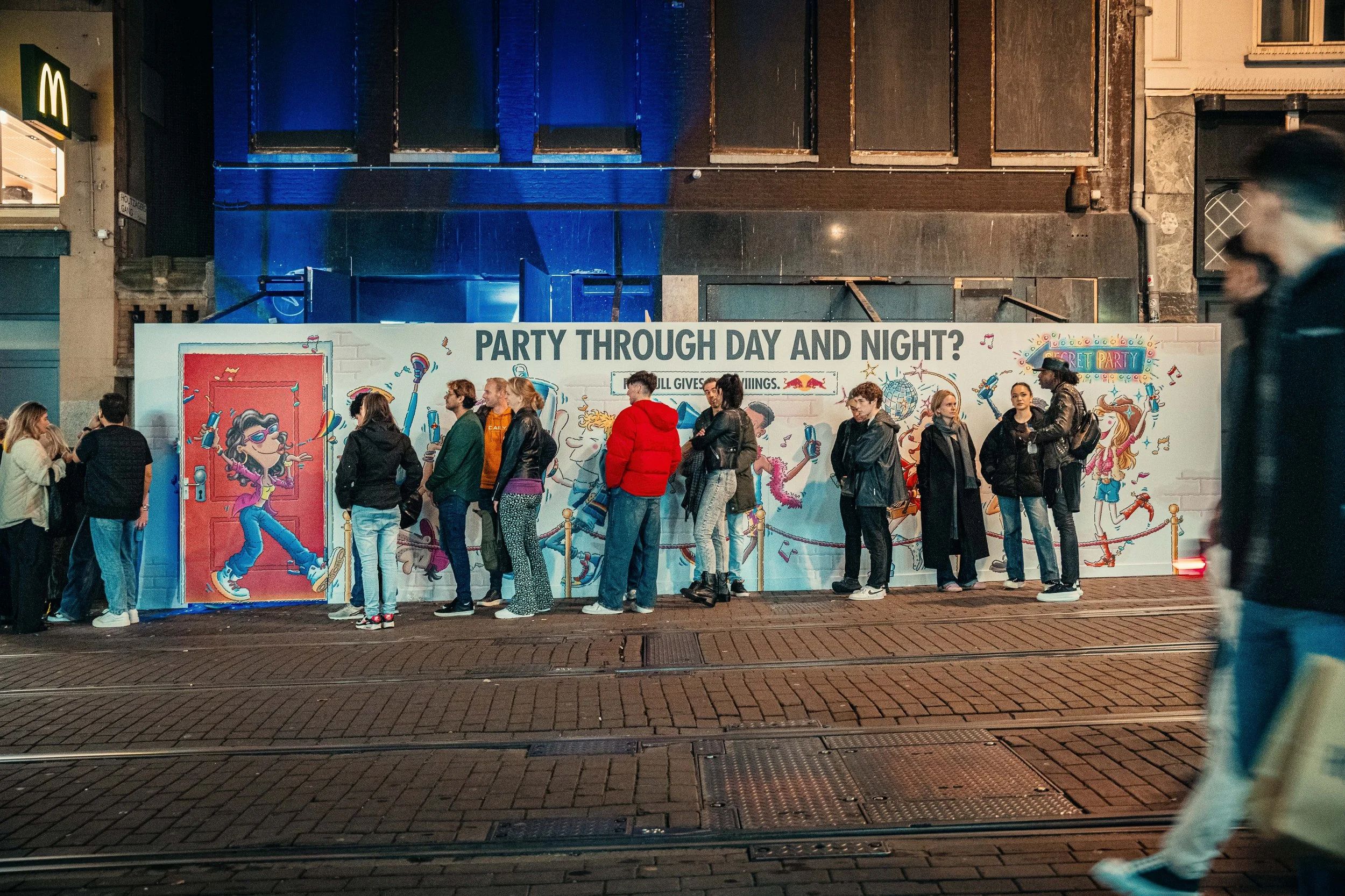 Een rij mensen wacht buiten een muur met kleurrijke illustraties en de tekst 'PARTY THROUGH DAY AND NIGHT?' onder een stadswand. Mensen staan in de rij, sommige chatten, anderen wachten rustig.