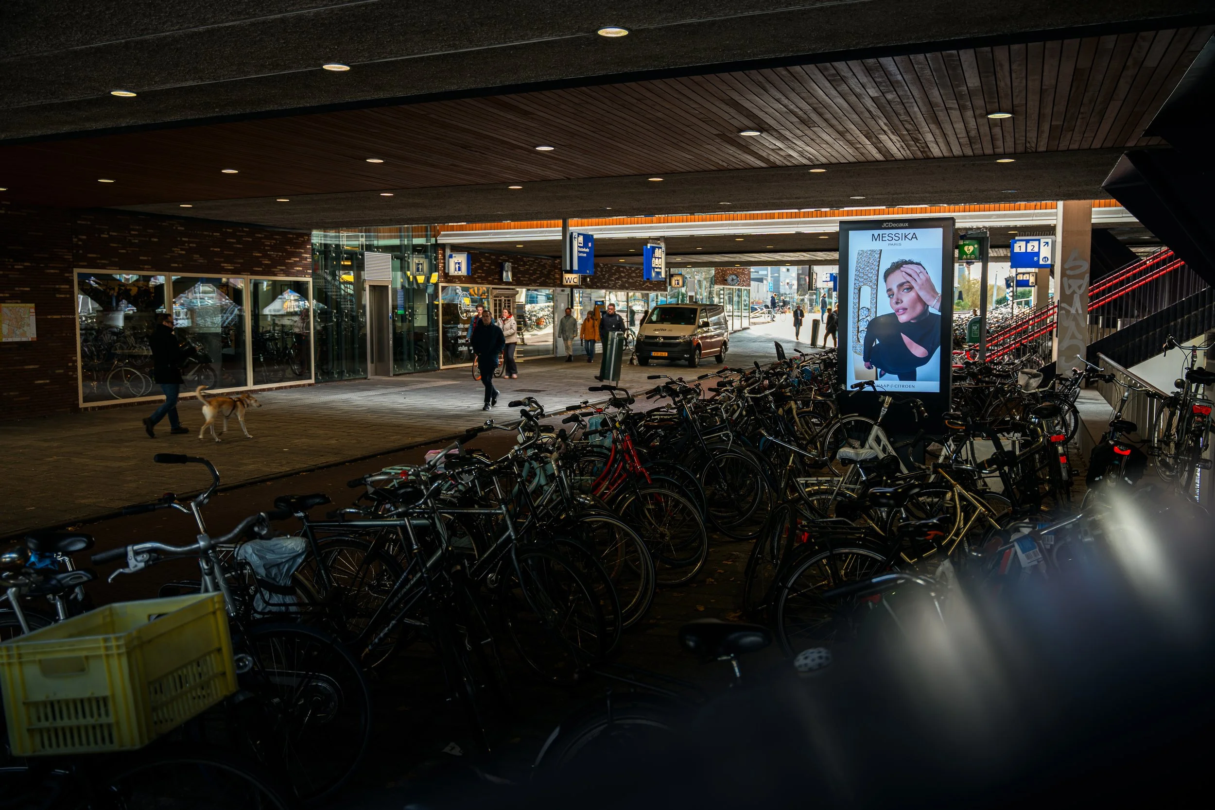 Overdekte fietsenstalling met rijen fietsen onder een brug of overkapping. Op de achtergrond mensen die lopen bij een gebouw met een digitale reclamezuil.